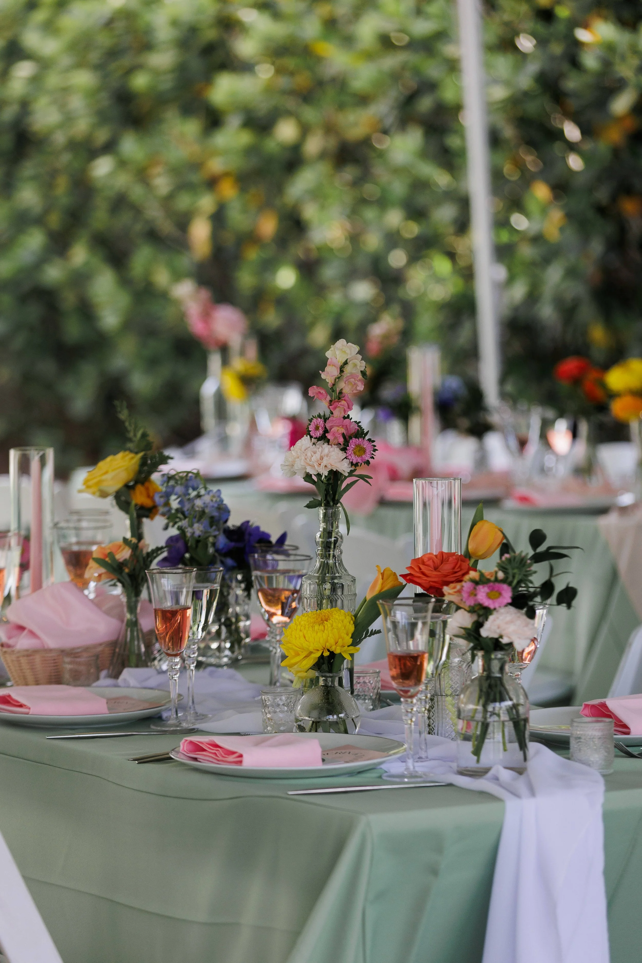 A beautifully decorated outdoor table set for a celebration with pink, yellow, orange, and purple flowers in glass vases, pink napkins, and pink drinks in wine glasses on a green tablecloth.