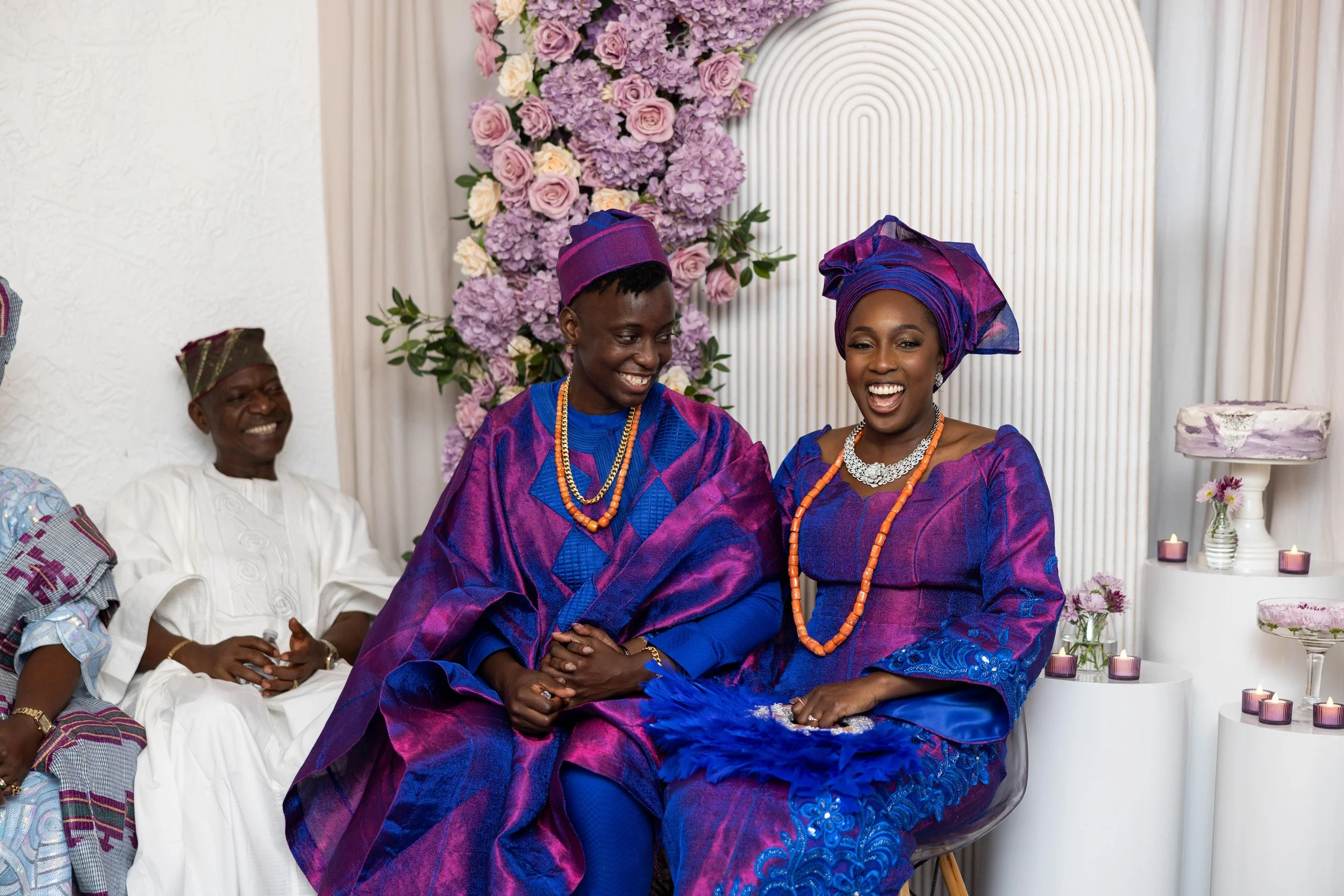 Two women in traditional Nigerian attire sitting and holding hands, smiling at each other during a celebration. A man dressed in white traditional clothing is seated behind them, also smiling. The background is decorated with pink and purple flowers 