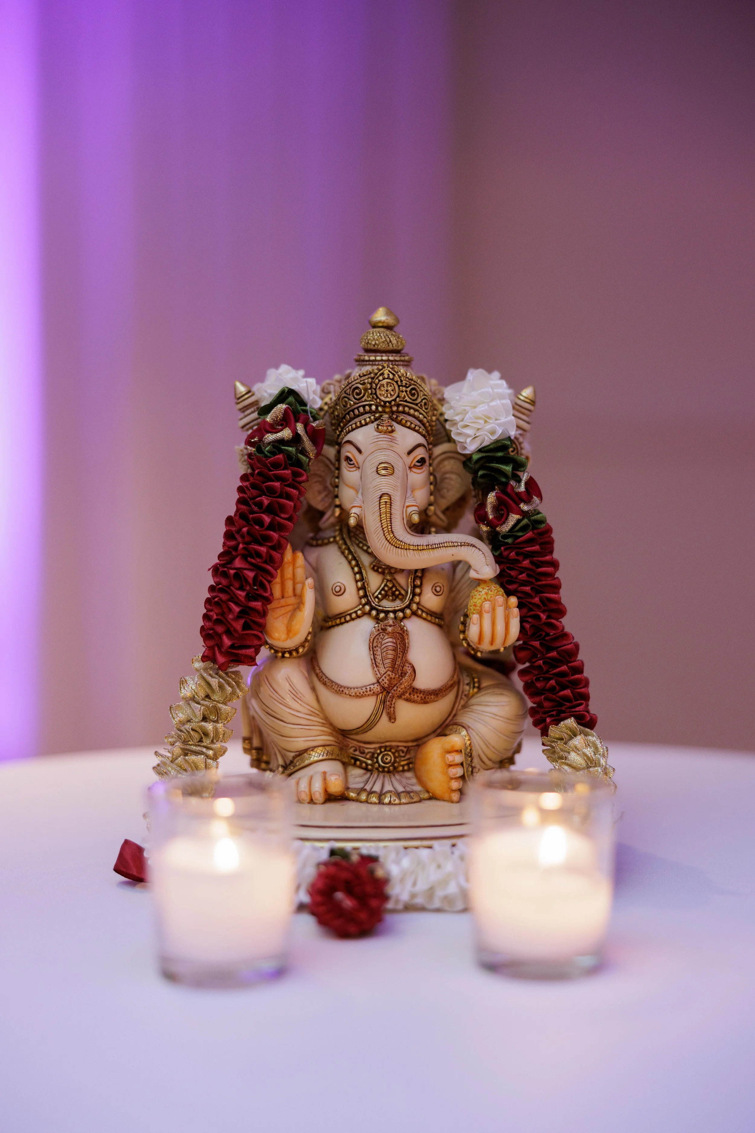 Statue of Lord Ganesha adorned with flower garlands, surrounded by lit candles on a white table.