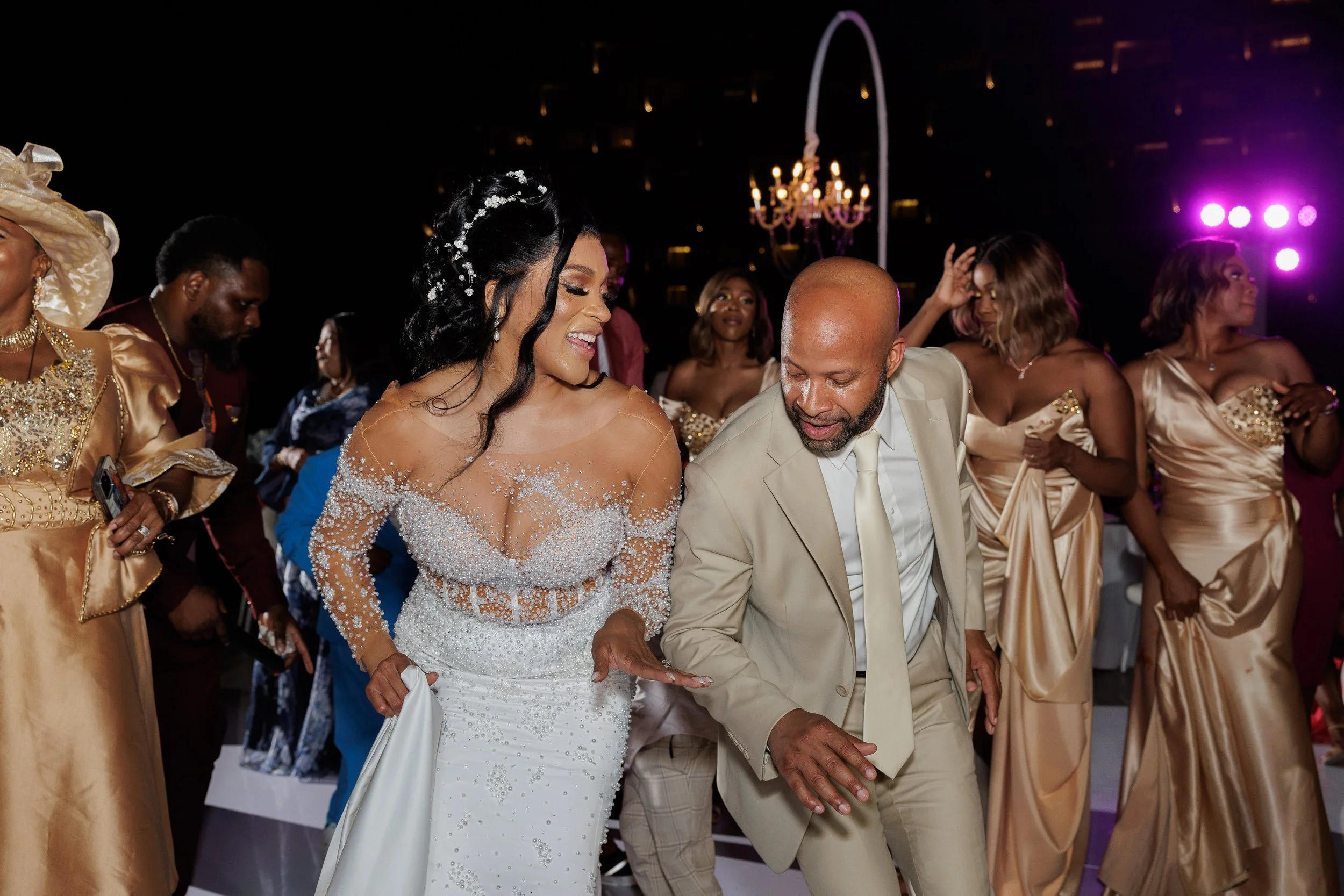 Couple dancing at a wedding reception surrounded by bridesmaids and guests at night with colorful lighting.
