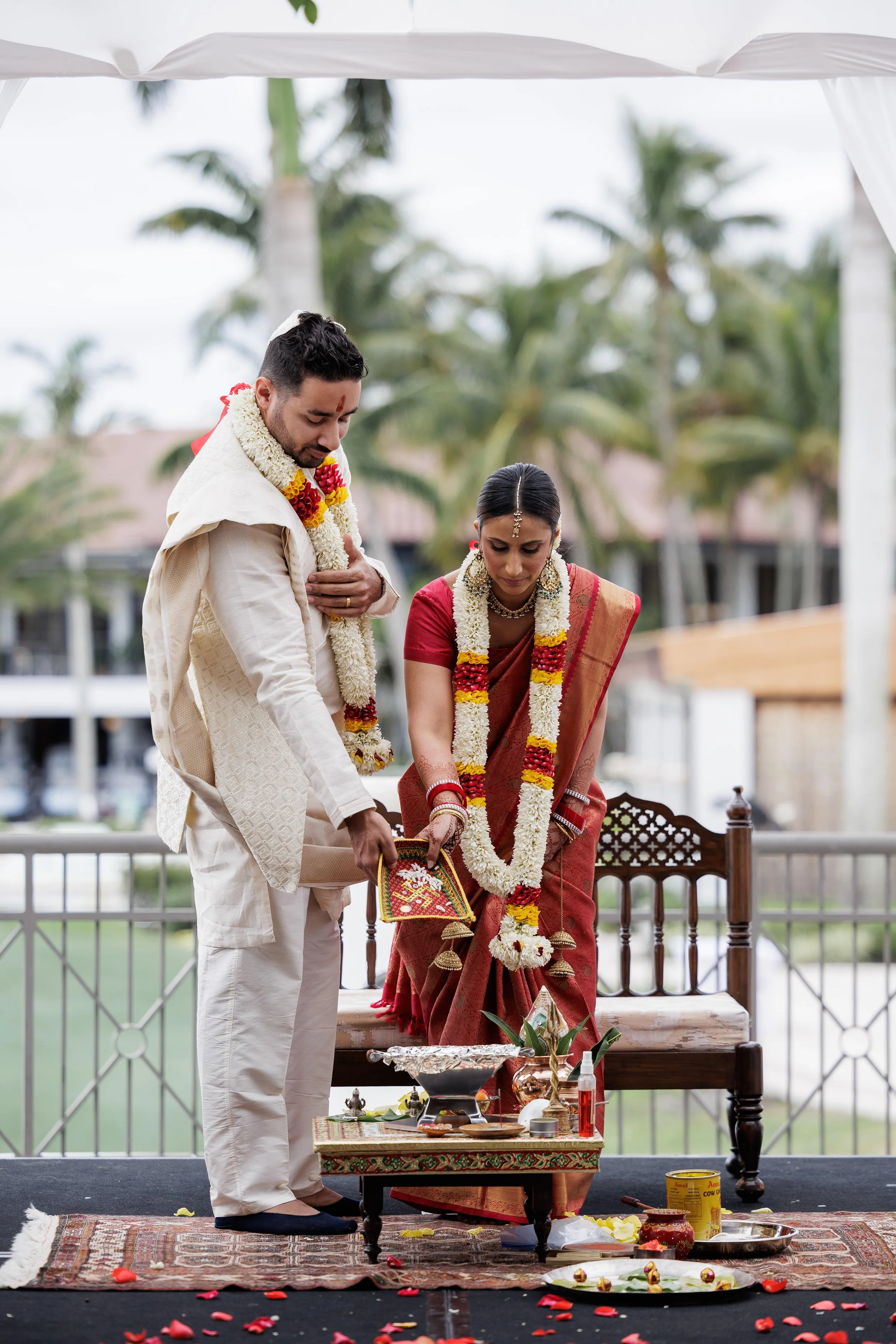 A couple dressed in traditional Indian wedding attire participating in a ceremonial ritual outdoors under a canopy, with palm trees and a building in the background.