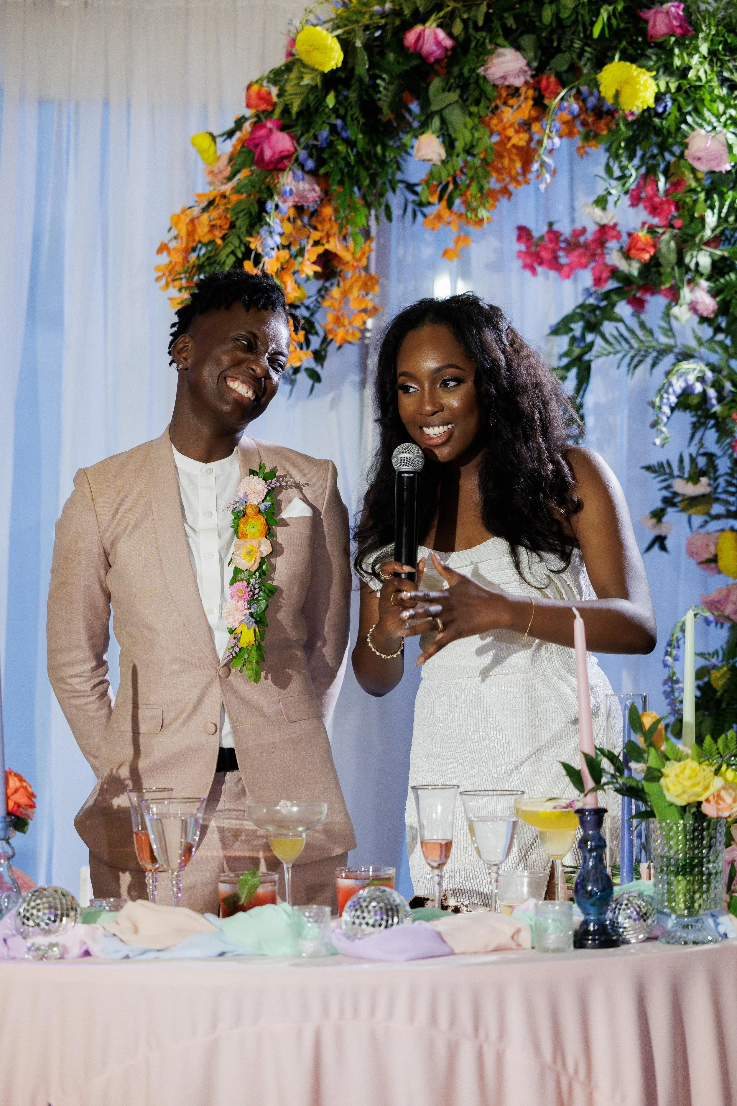 Two women at a wedding reception, one in a tan suit with a floral corsage, the other in a white dress with a microphone, standing in front of a colorful floral arch.