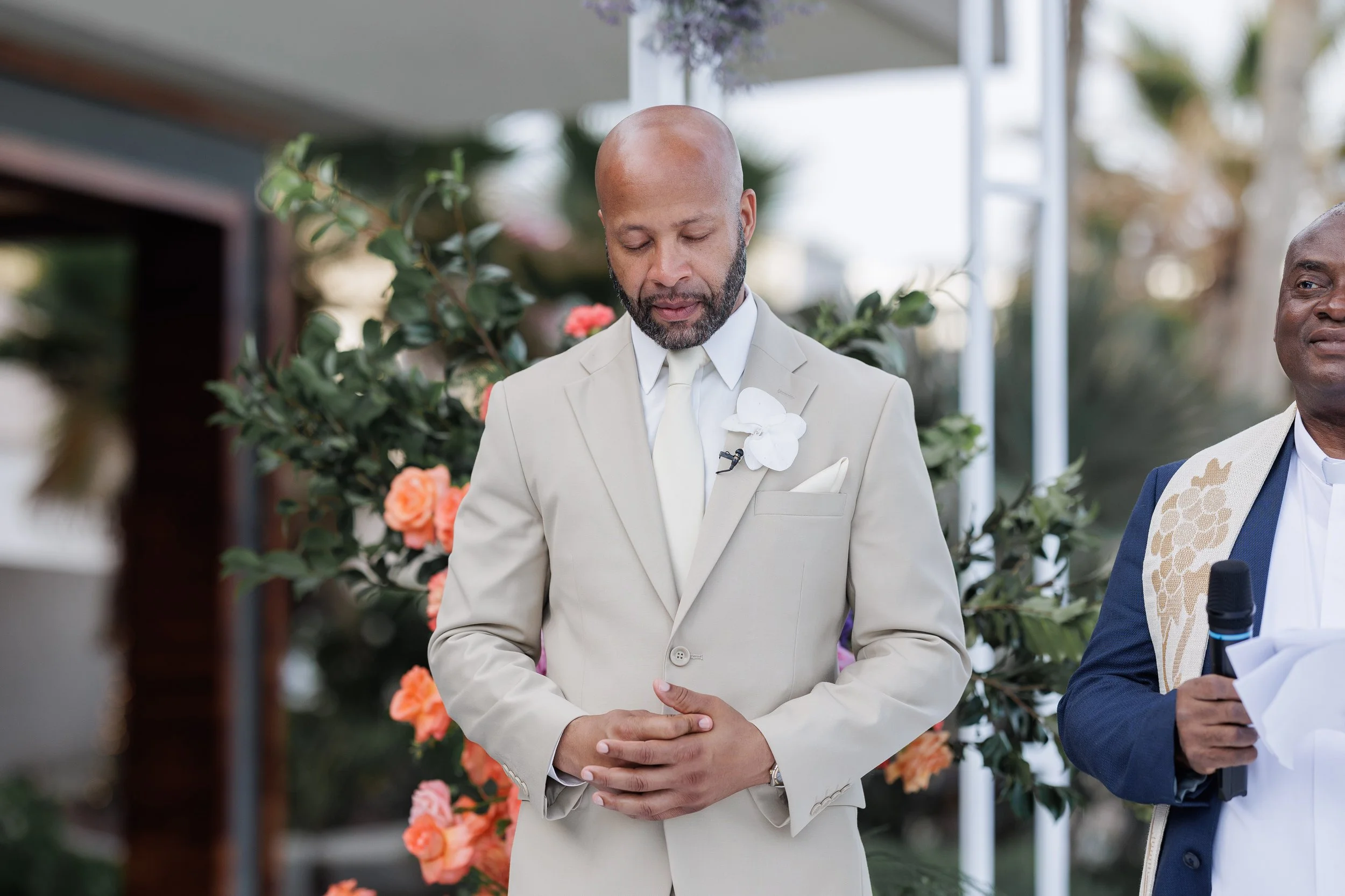 A man in a beige suit with a white tie and floral boutonniere, standing with his hands clasped during a ceremony. Another man in a navy suit and white shirt holding a microphone is partially visible on the right. Background has greenery and blooming 