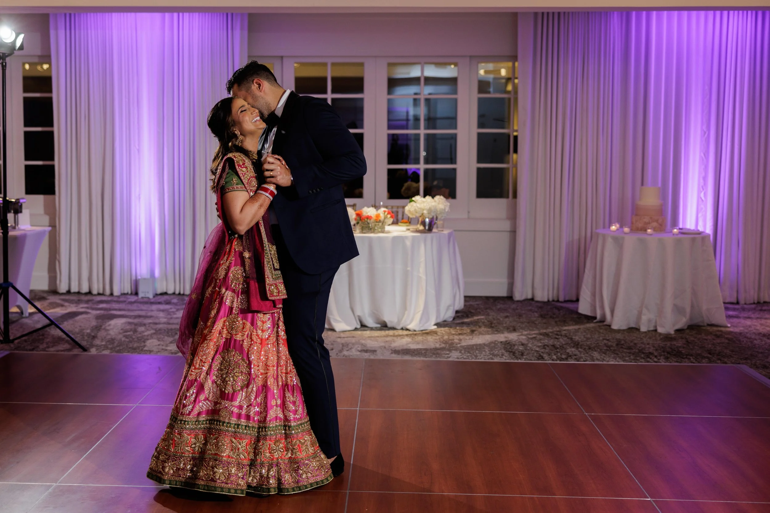 A couple dancing at a wedding reception, with the woman dressed in a traditional Indian bridal outfit and the man in a dark suit. They are inside a decorated hall with purple lighting, white curtains, and tables with floral arrangements and a wedding