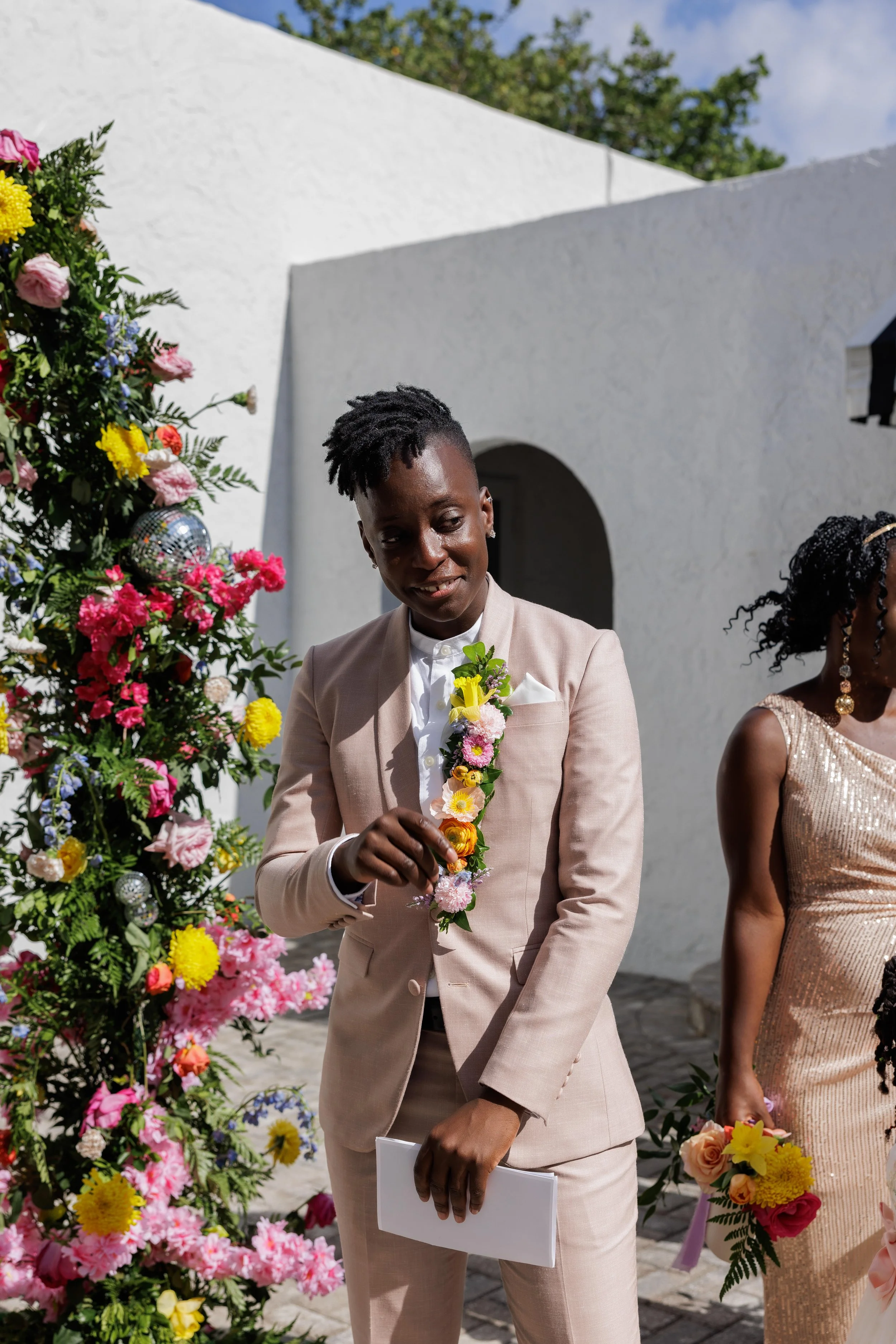 Person in light pink suit at outdoor wedding, decorated with colorful flowers and greenery.