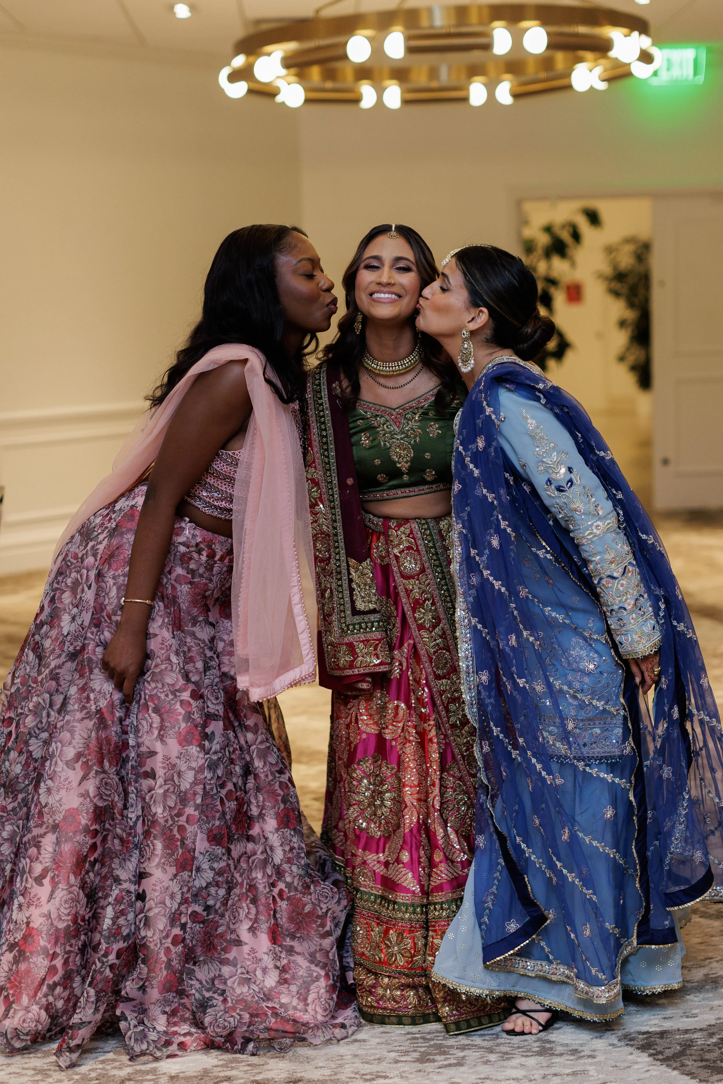 Three women dressed in colorful traditional Indian attire sharing a kiss on the cheeks and smiling, in an indoor setting with elegant lighting and decor.