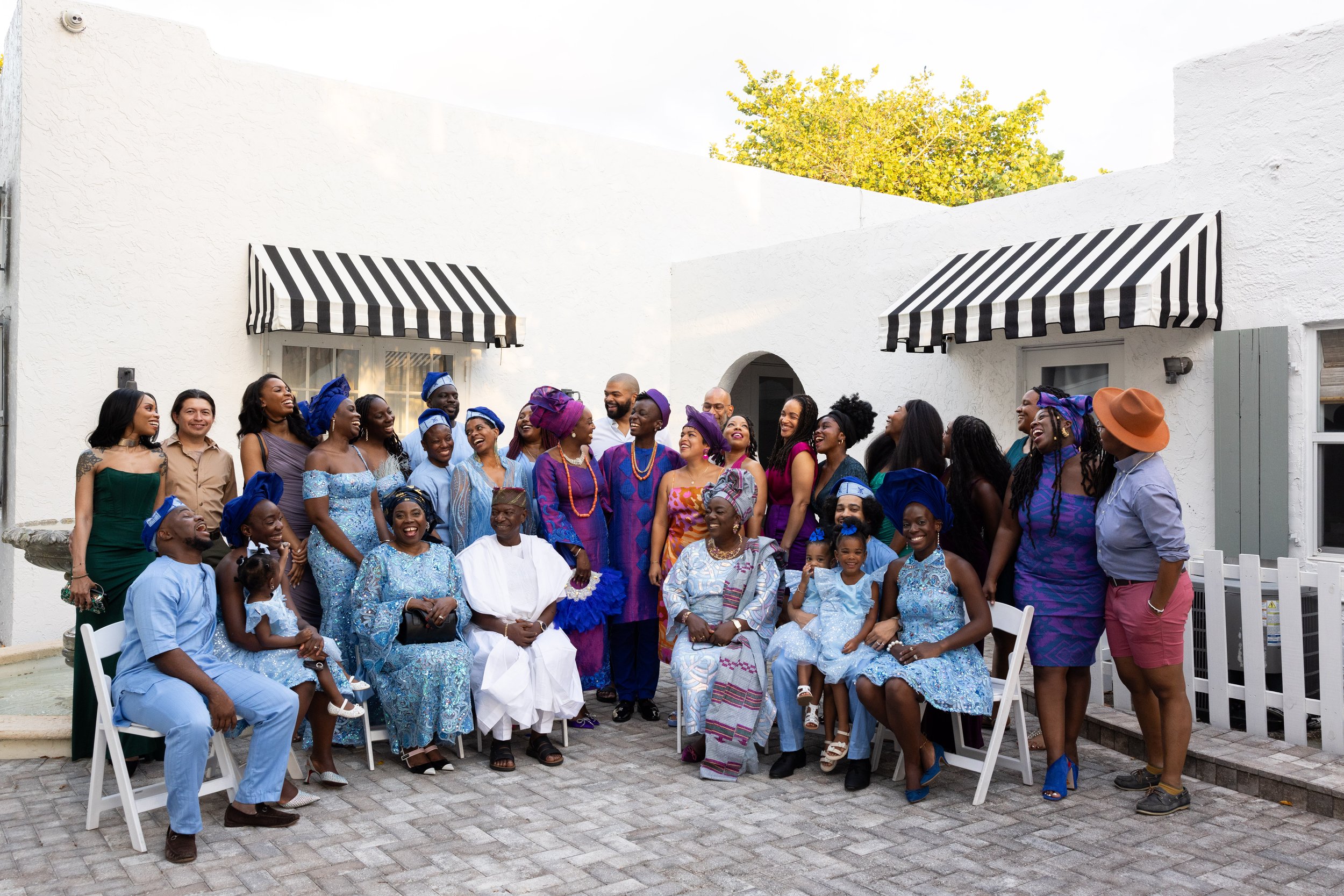 A large group of people, including children and adults, gathered outside in front of white buildings with striped awnings, celebrating and smiling, dressed in colorful traditional and modern clothing, with some women wearing headwraps and men in trad