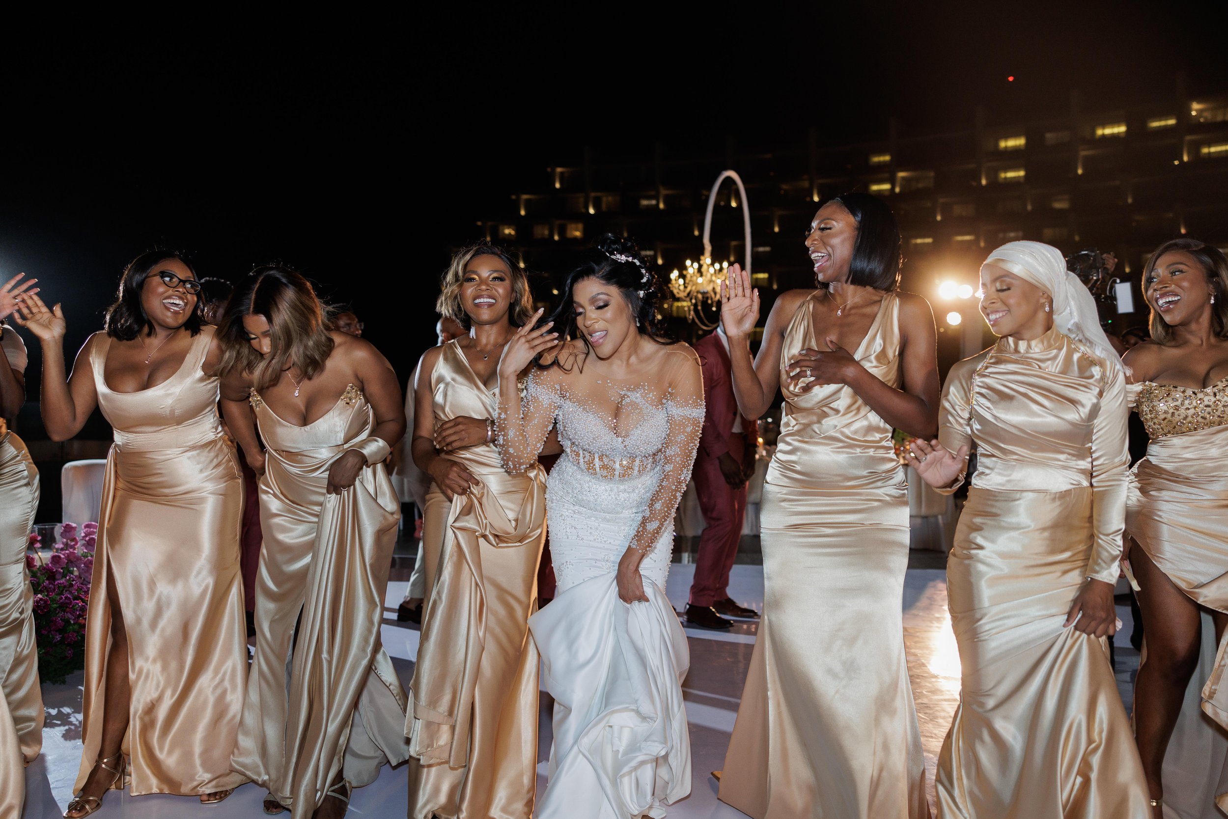 A group of women dancing at a wedding celebration at night, with a city skyline and the Gateway Arch illuminated in the background.