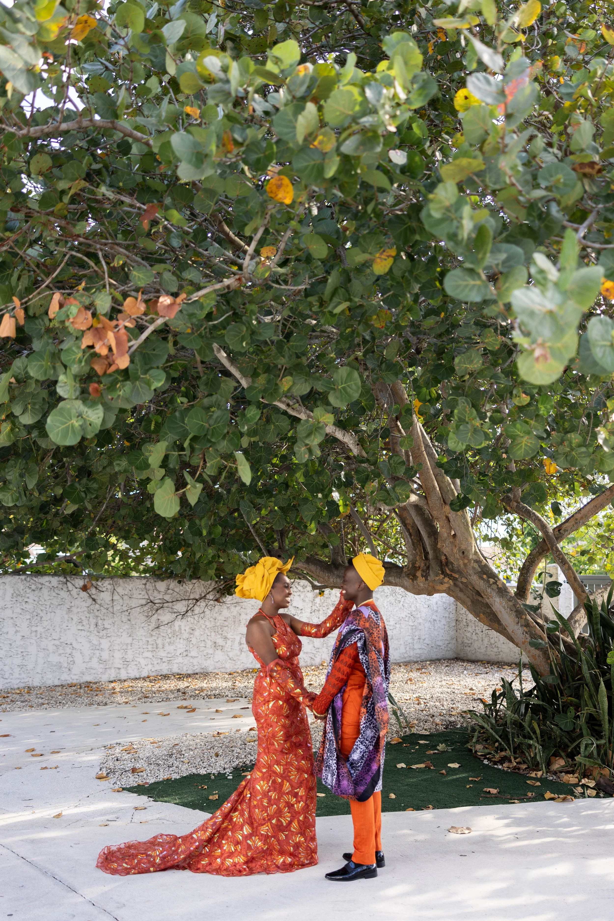 Two people dressed in colorful traditional African attire, holding hands and smiling at each other under a large leafy tree.