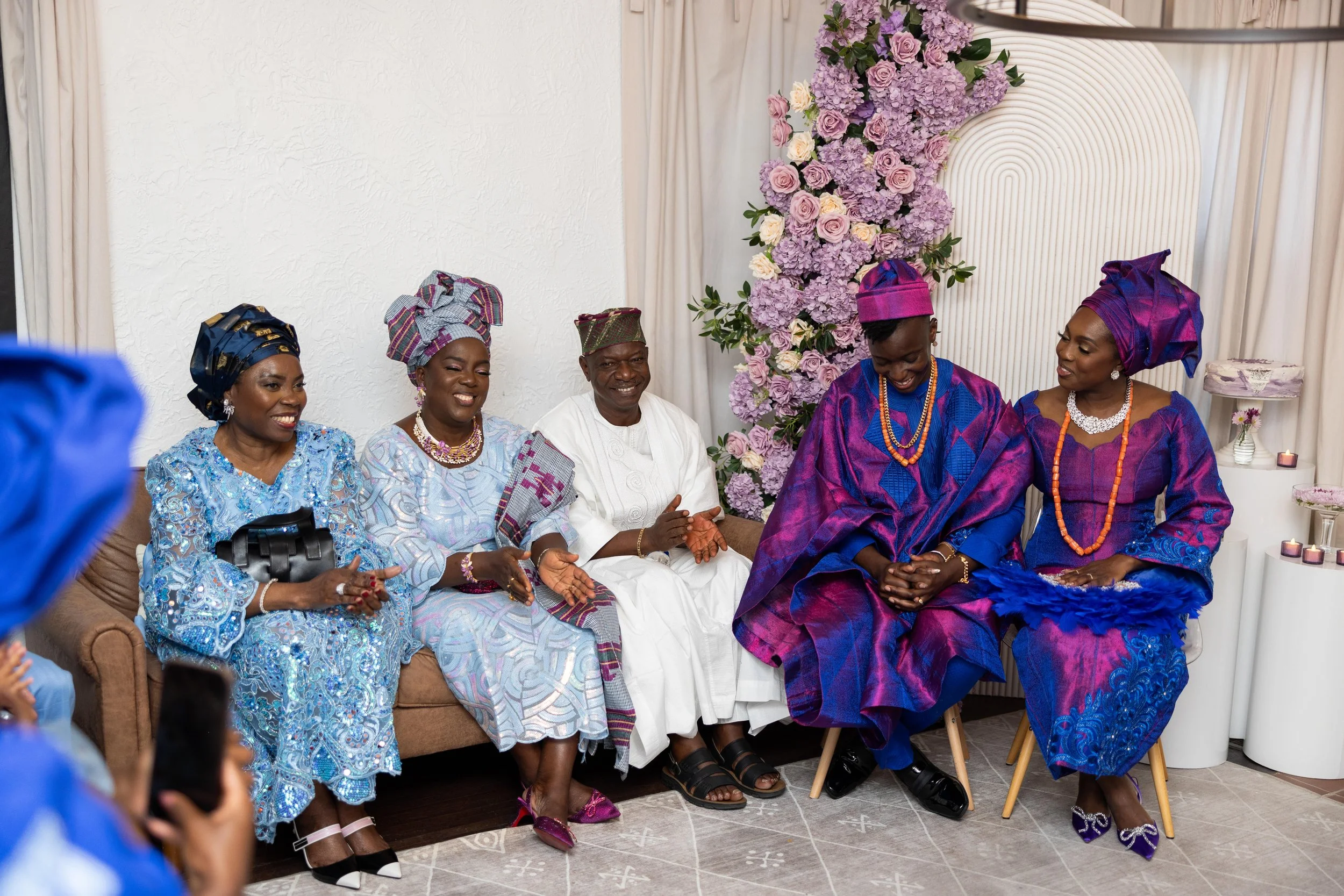 Six people sitting on a sofa, dressed in traditional Nigerian attire, attending a celebration with floral decorations.