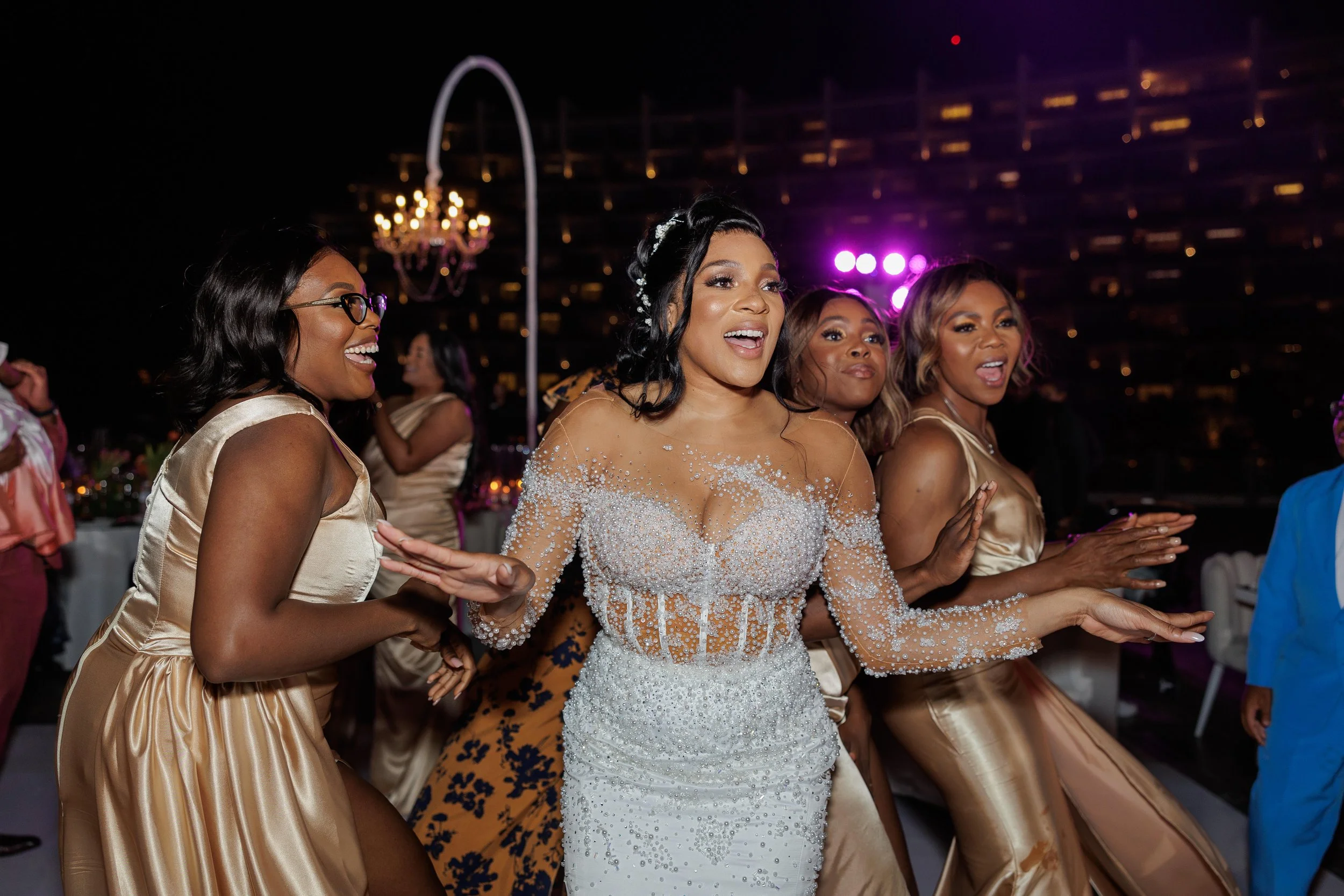 Women dancing and celebrating at a wedding reception, dressed in elegant wedding attire with the bride in a beaded gown, on a rooftop at night with city lights and a chandelier in the background.