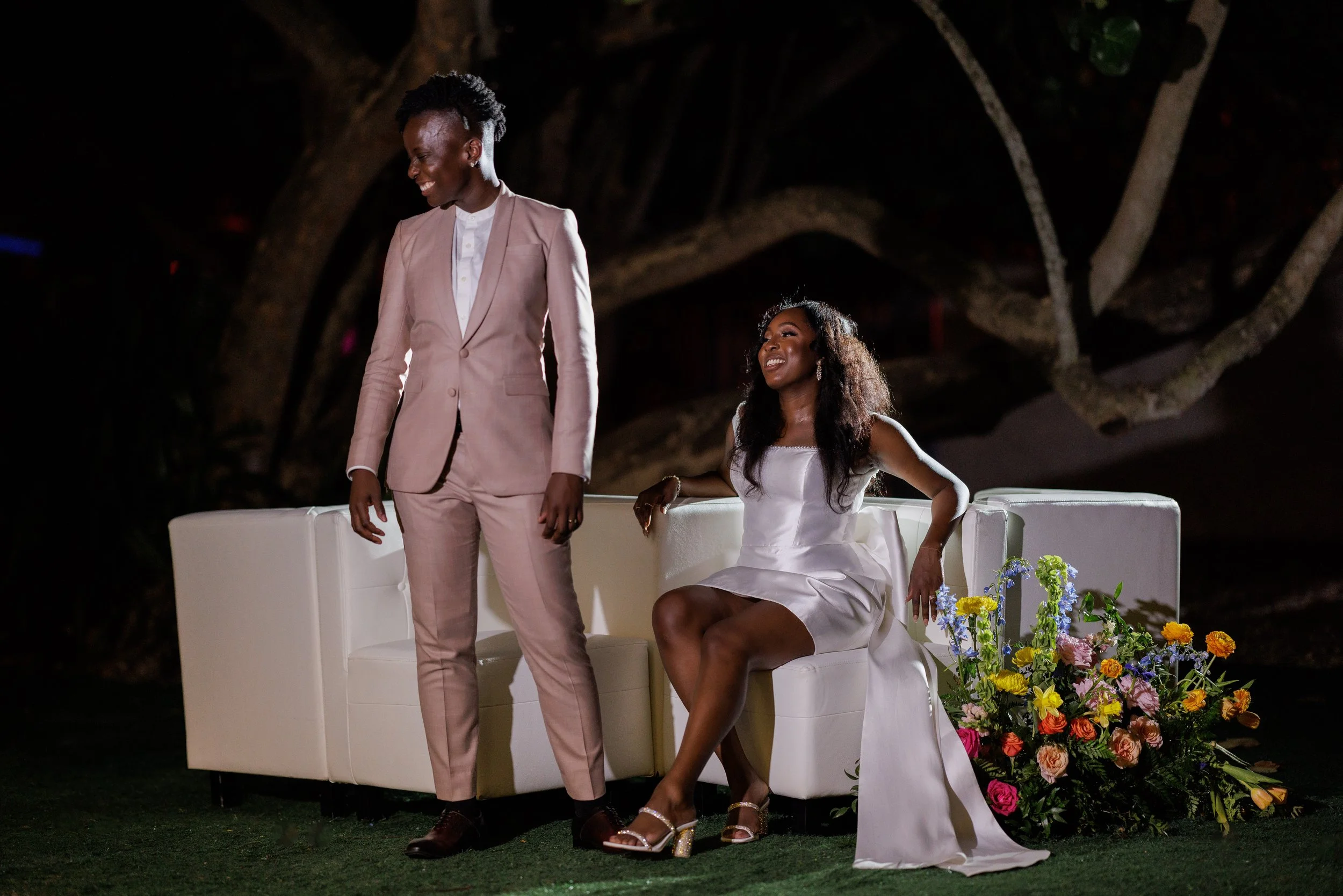 A woman in a white dress sitting on a sofa next to a man in a light pink suit at an outdoor event with a floral arrangement nearby.