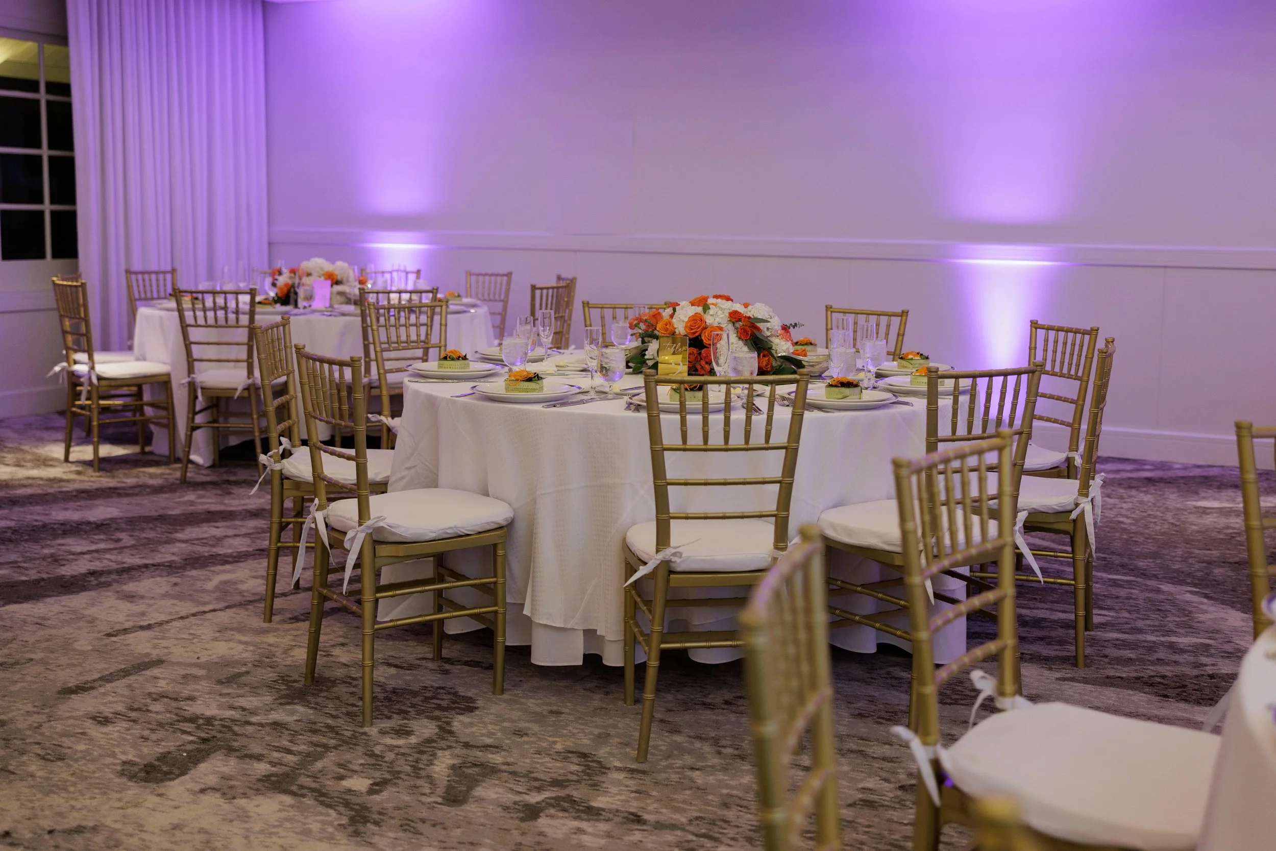Elegant banquet hall with round tables draped in white tablecloths, decorated with floral centerpieces, surrounded by gold chiavari chairs with white cushions, and purple uplighting on the walls.