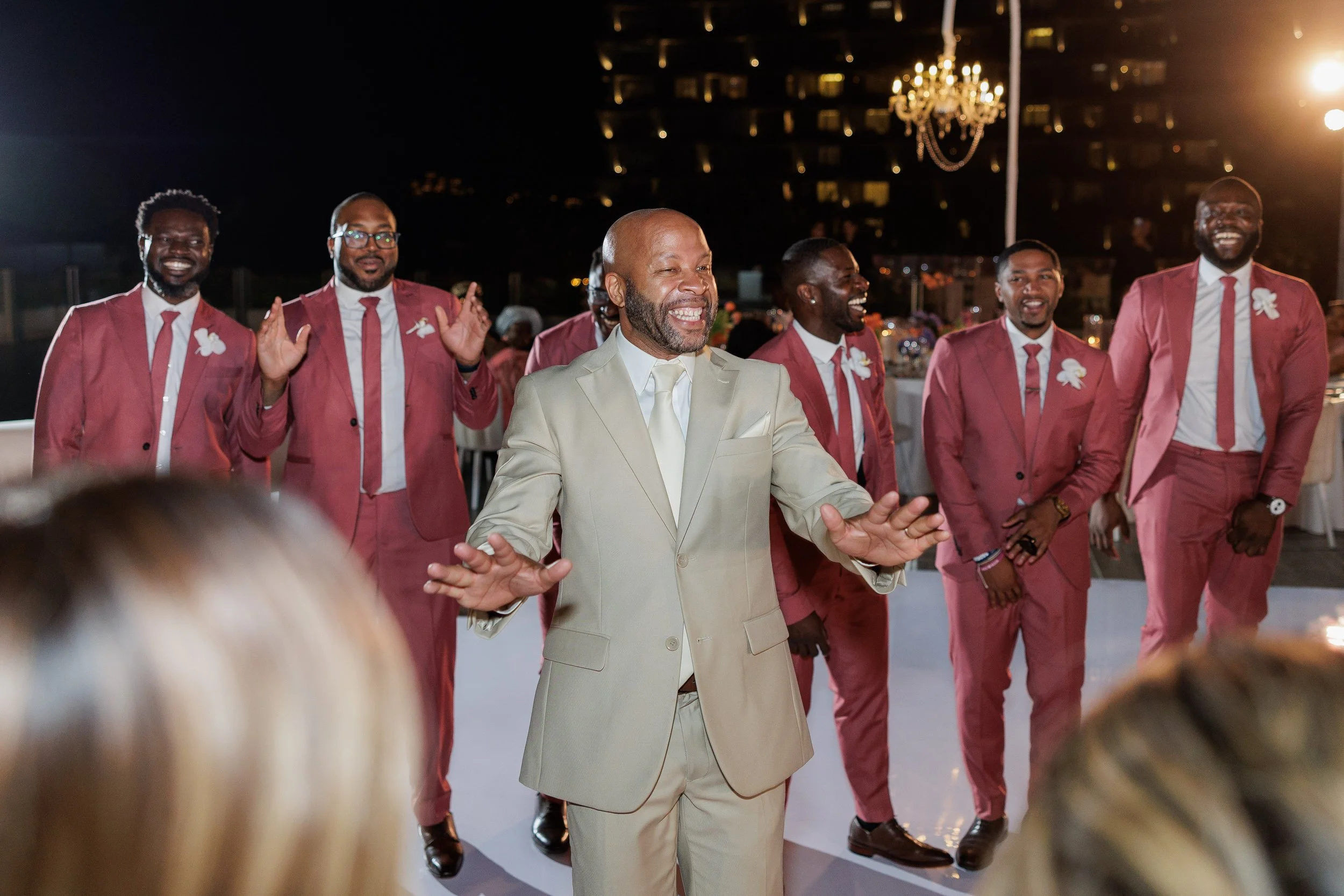 Man in a light beige suit smiling and dancing at a celebration with a group of men in matching pink suits, outside at night with a lit chandelier and city lights in the background.