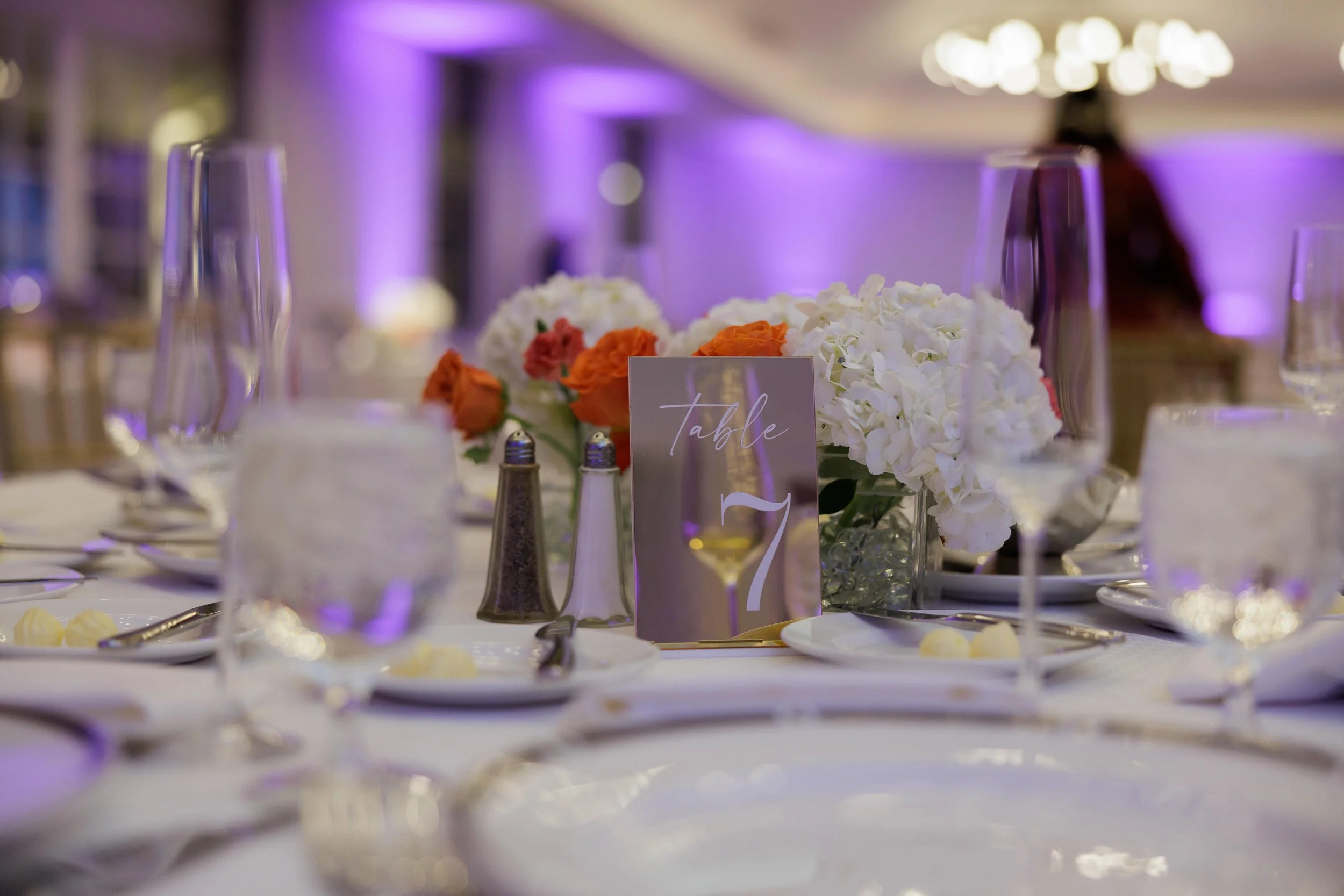 Elegant banquet table setting with floral centerpiece, wine glasses, salt and pepper shakers, and a table number card for table 7, in a decorated banquet hall with purple lighting.