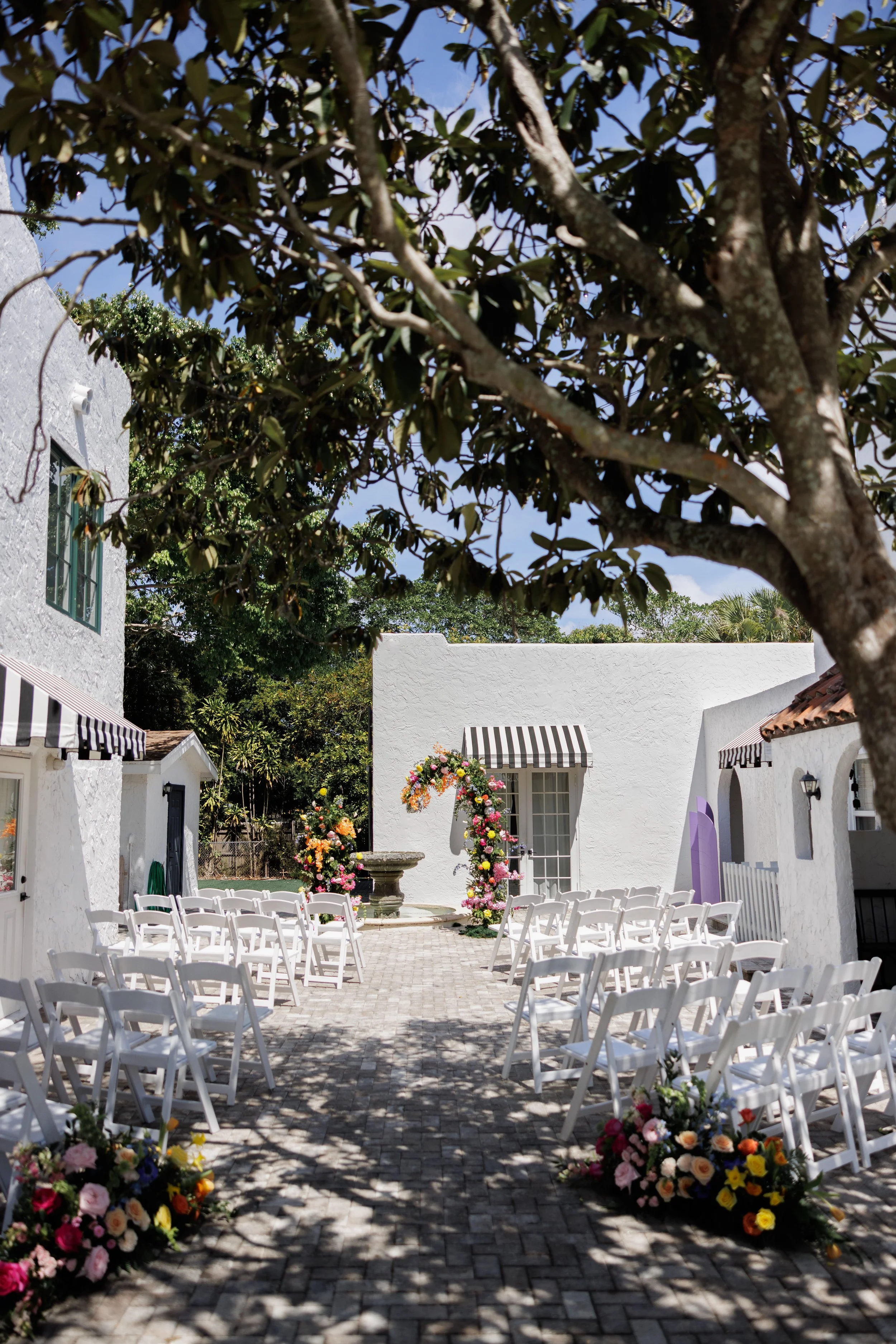 Outdoor wedding ceremony setup with white folding chairs arranged on brick pavement, decorated with colorful flower arrangements, in front of a white building with a floral arch and fountain, shaded by large trees.