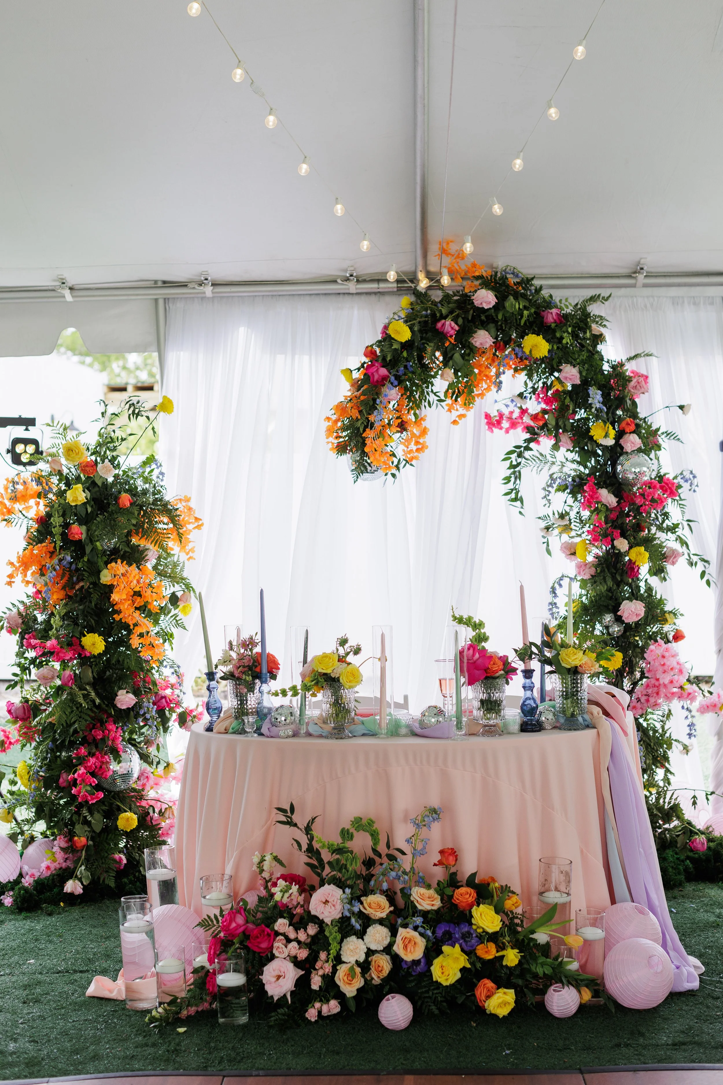 A decorated event table with a floral arch above, surrounded by colorful floral arrangements, candles, and assorted decorations in a white tent.