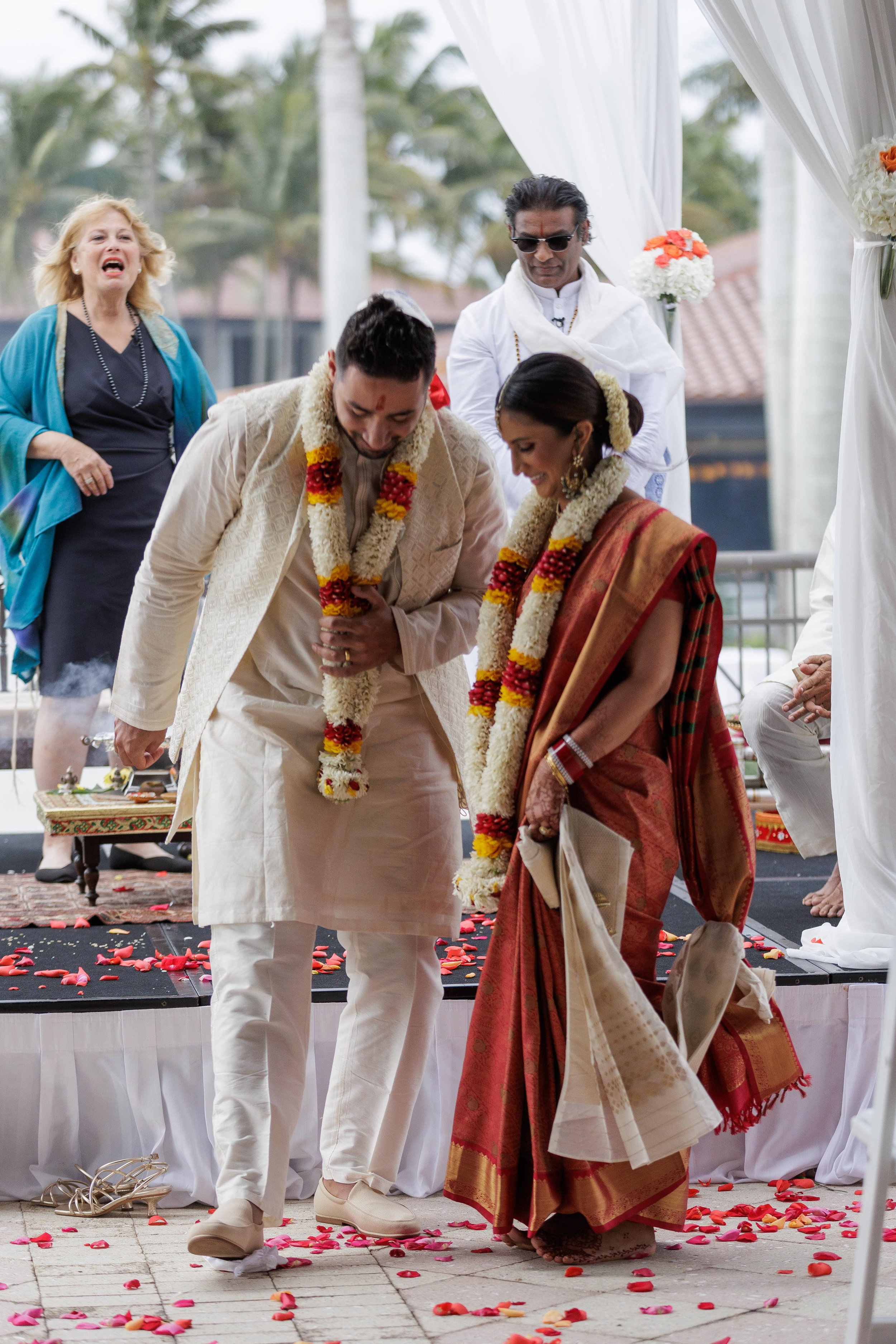 A traditional Indian wedding ceremony with bride and groom, dressed in traditional attire and wearing flower garlands, bowing to each other, surrounded by other participants and flower petals on the ground.