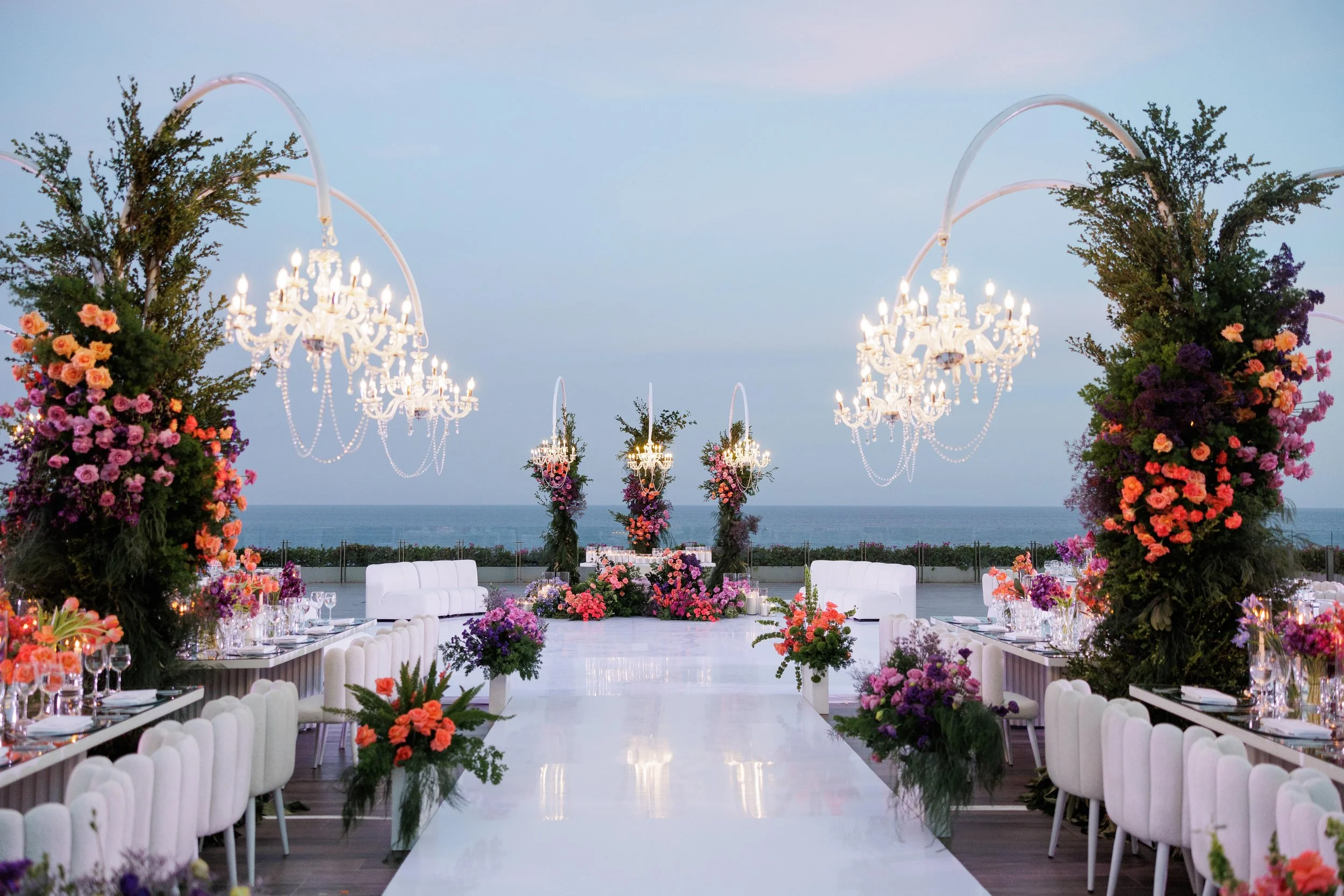 Elegant outdoor wedding reception setup on a terrace overlooking the ocean, decorated with large floral arrangements, chandeliers, and seating on both sides of a white aisle.