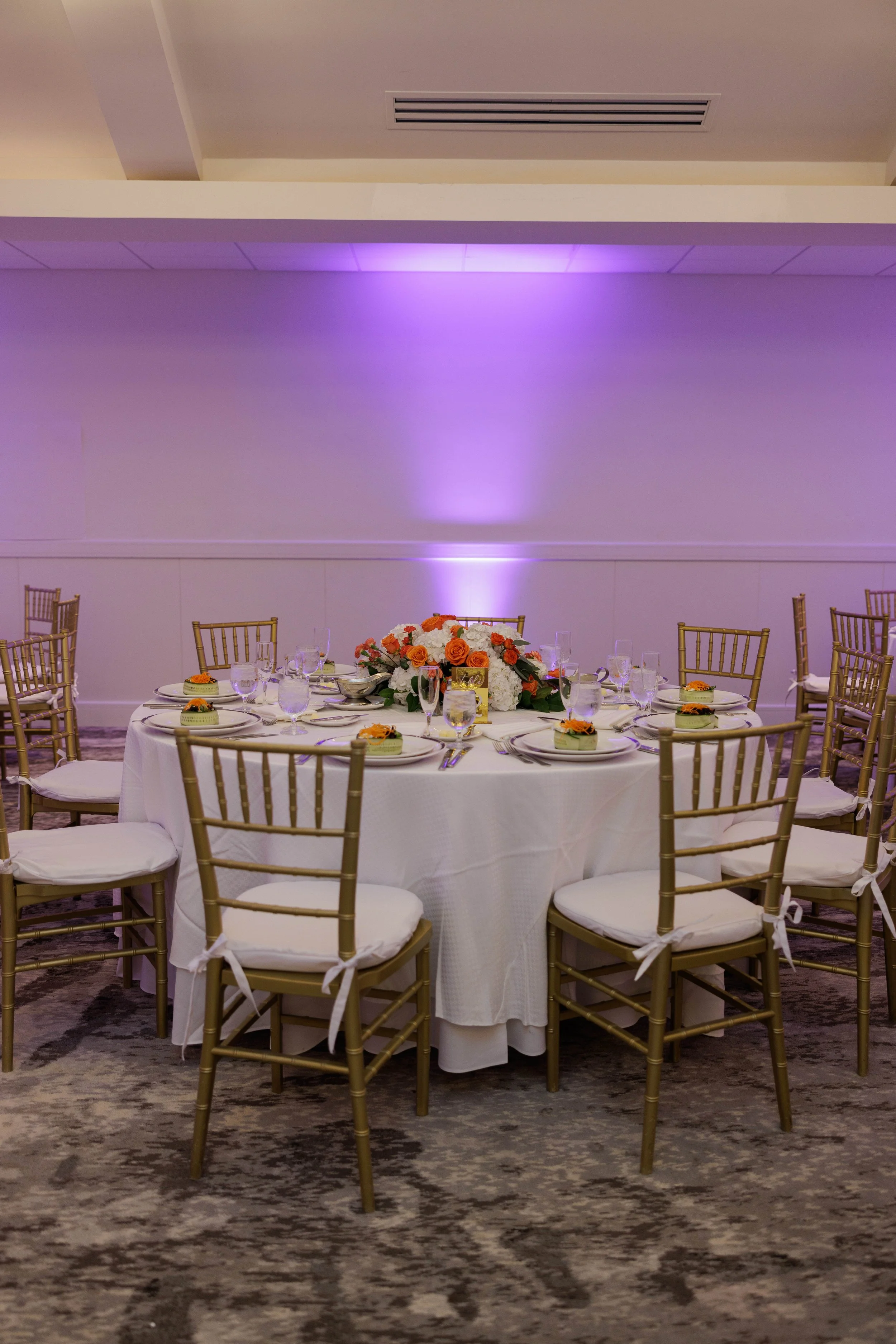 Elegant round banquet table decorated with a floral centerpiece, set with plates, wine glasses, and utensils, surrounded by gold-colored chairs with white cushions, in a banquet hall with purple lighting.