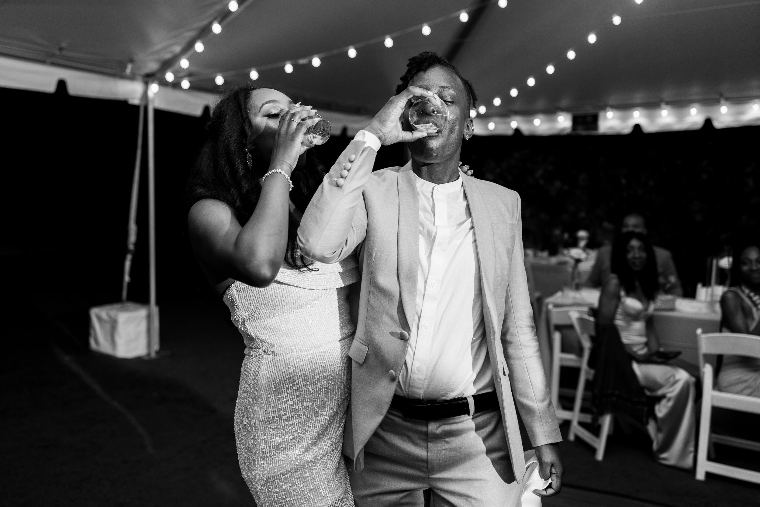 Black and white photo of a man and woman wearing formal attire, drinking from glasses at an outdoor event decorated with string lights, with seated guests in the background.