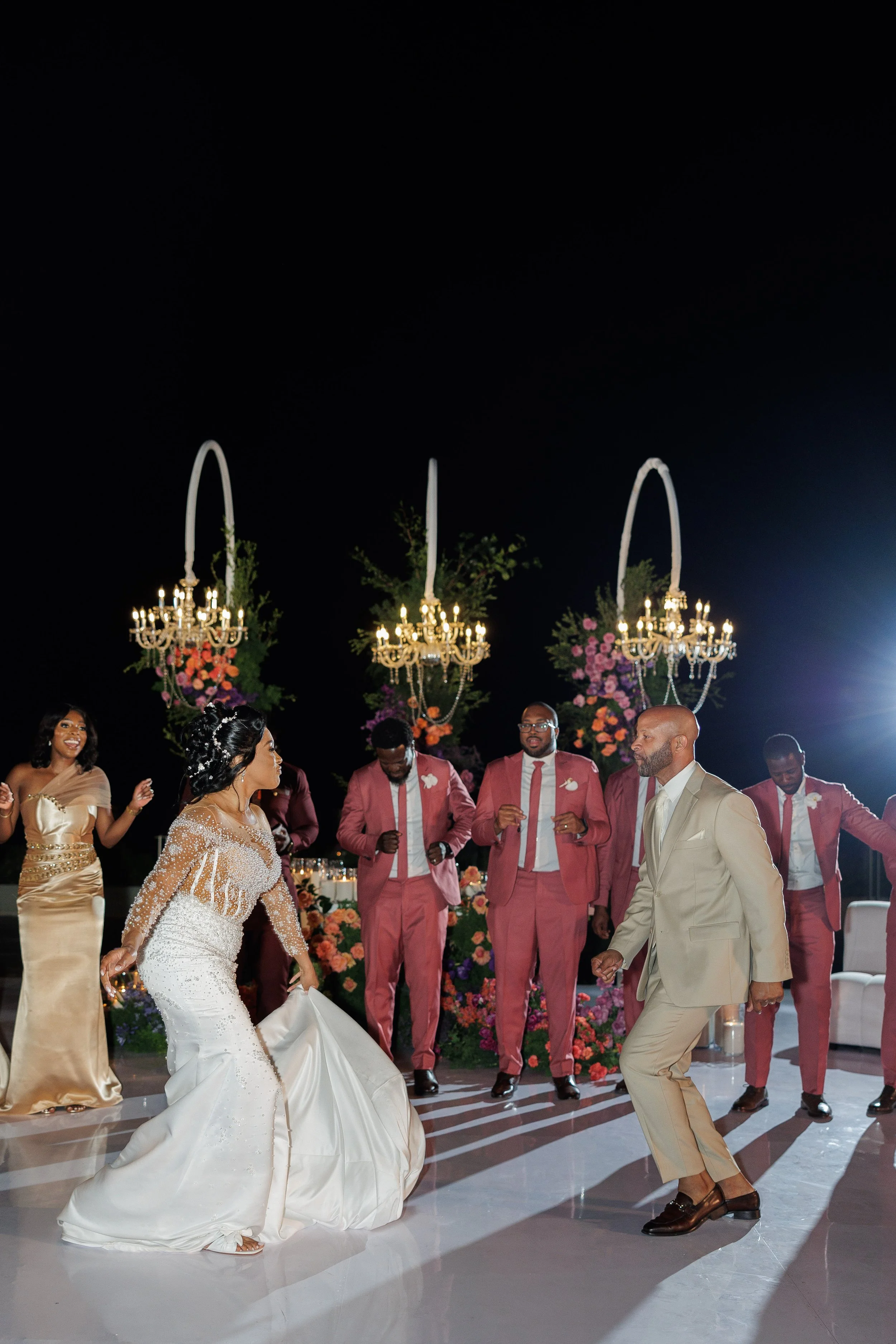 A bride and groom dancing at their wedding reception with guests dancing around them and floral decorations in the background.