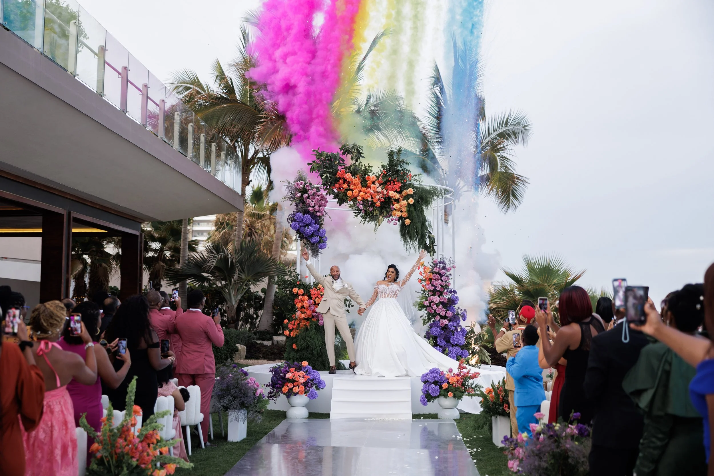 A wedding ceremony outdoors with a bride and groom celebrating under a floral arch as colorful smoke and powder are released above them. Guests are taking photos.