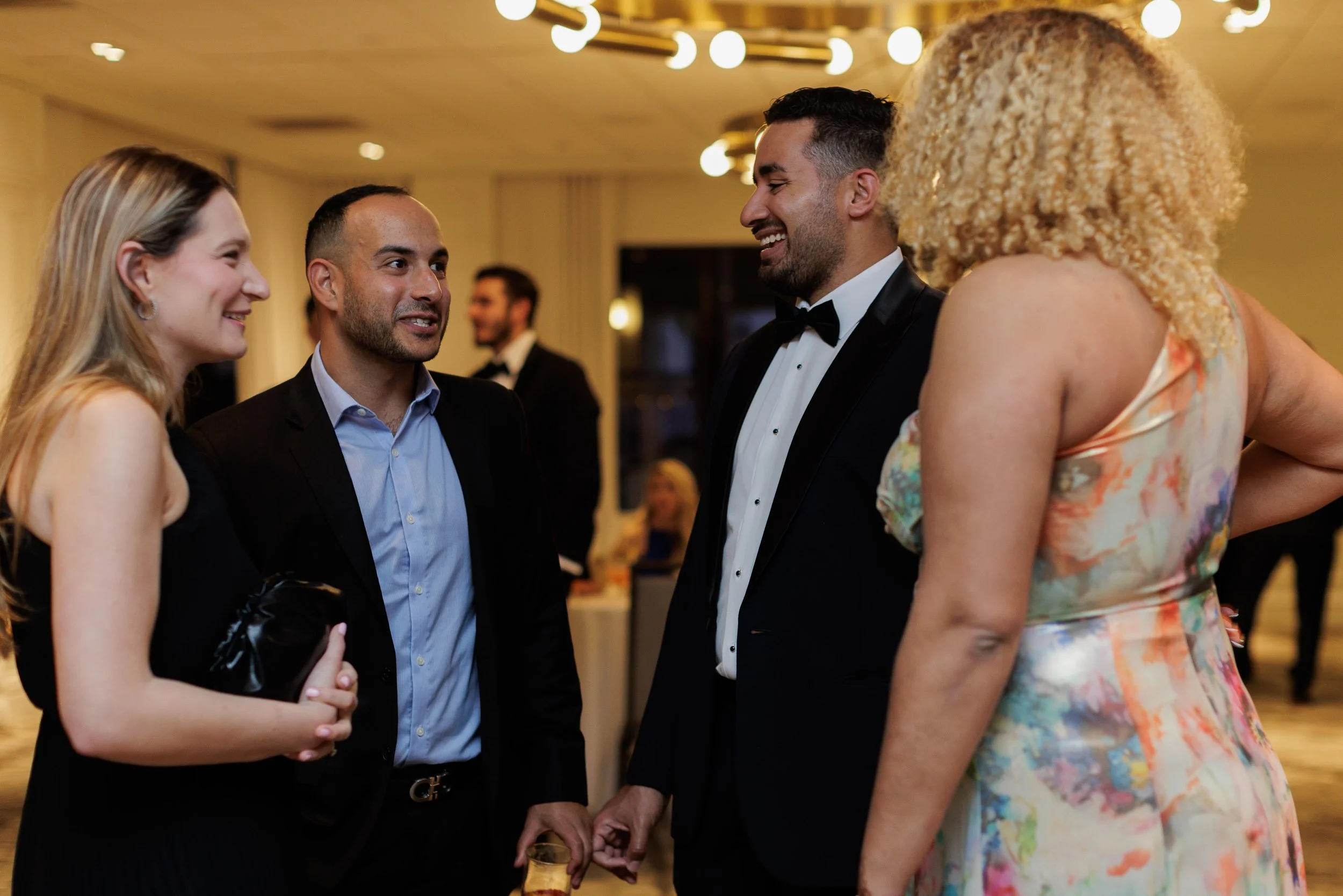 Group of four adults smiling and talking at a formal event in a well-lit room with hanging lights, with a man in a tuxedo and a woman in a floral dress.