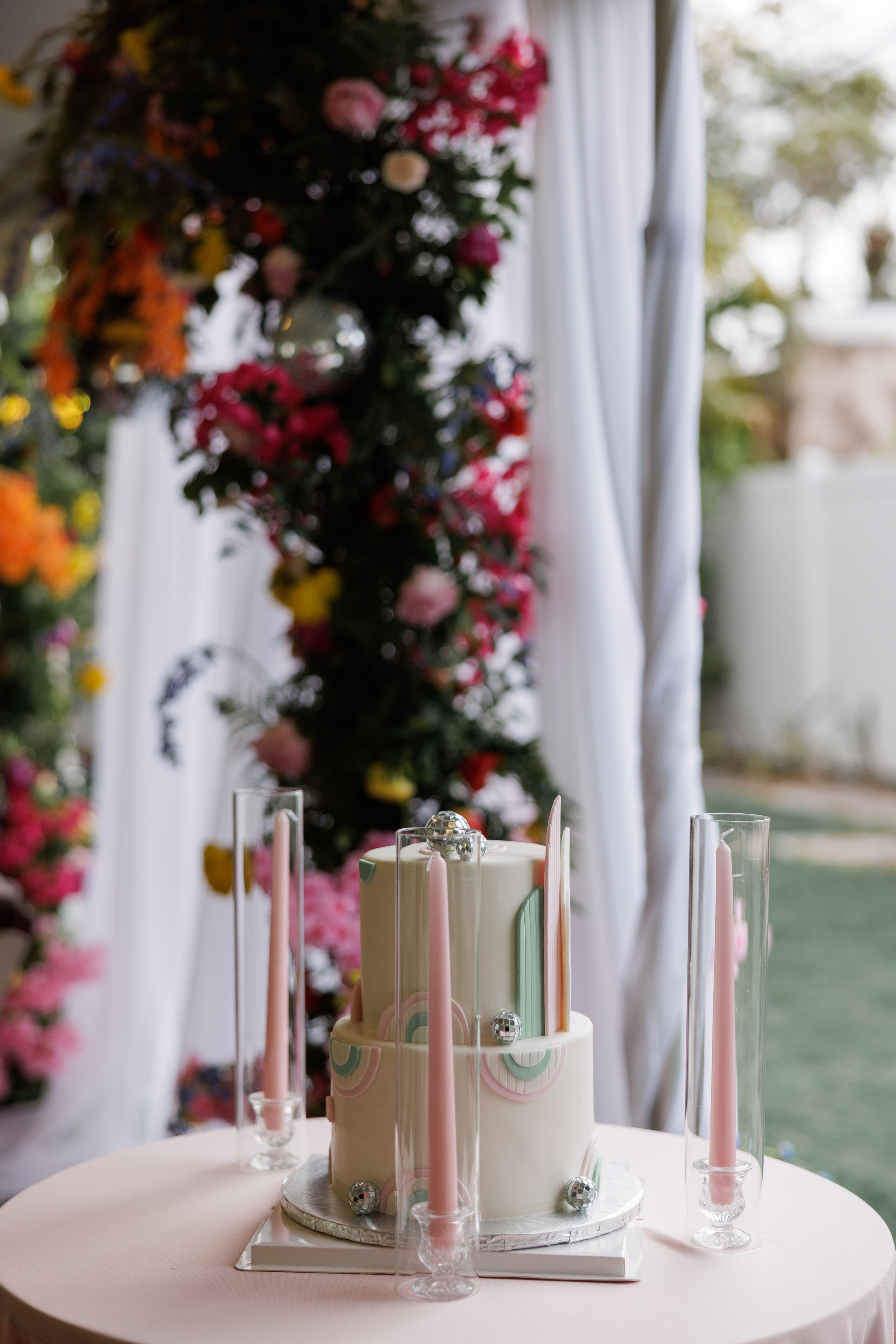 A two-tiered white wedding cake decorated with pastel rainbow designs and silver balls, surrounded by pink taper candles in glass holders, on a pink tablecloth with floral arrangements and a floral arch in the background.