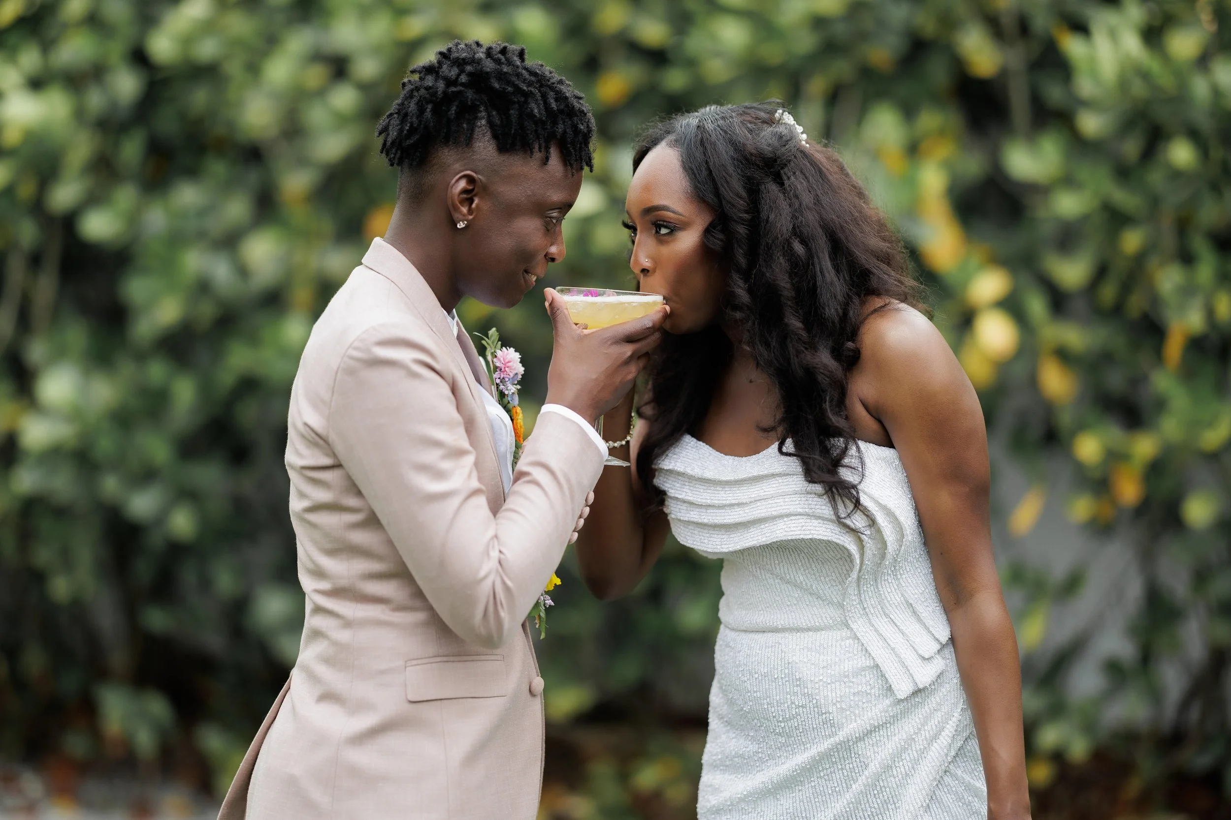A woman in a white dress is being served a cocktail by a man in a beige suit at an outdoor event, surrounded by green foliage.