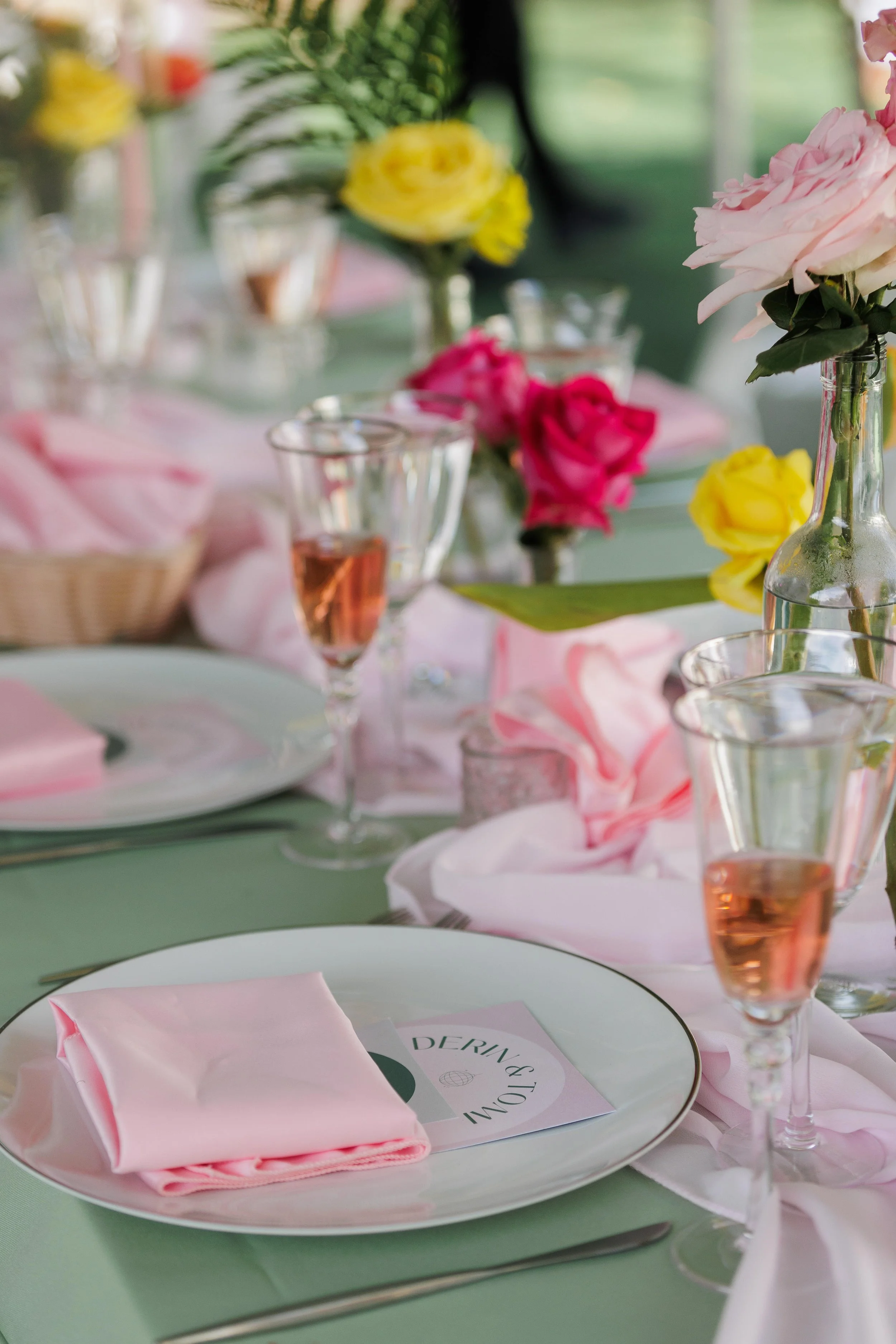 Elegant table setting with pink napkins, wine glasses with rosé, and floral centerpieces including pink, yellow, and white flowers.