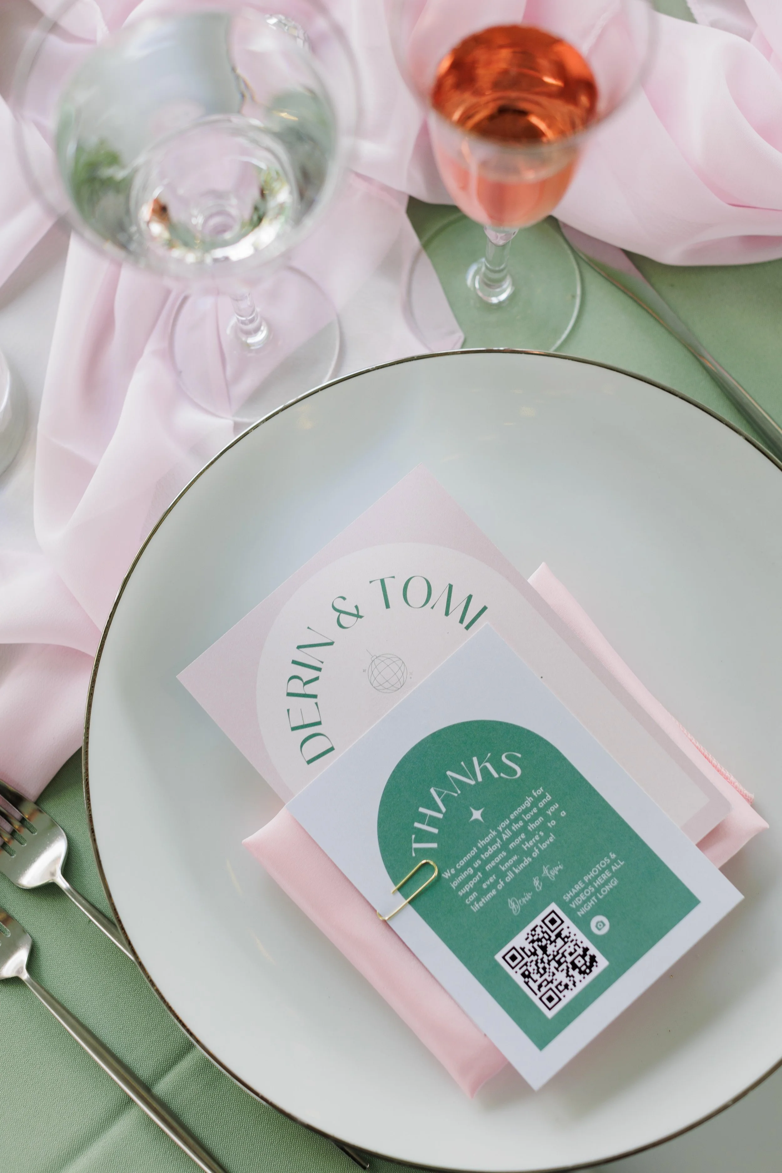Wedding reception table setting with a white plate, pink napkin, green place card, and pink and clear glasses of Champagne in the background.
