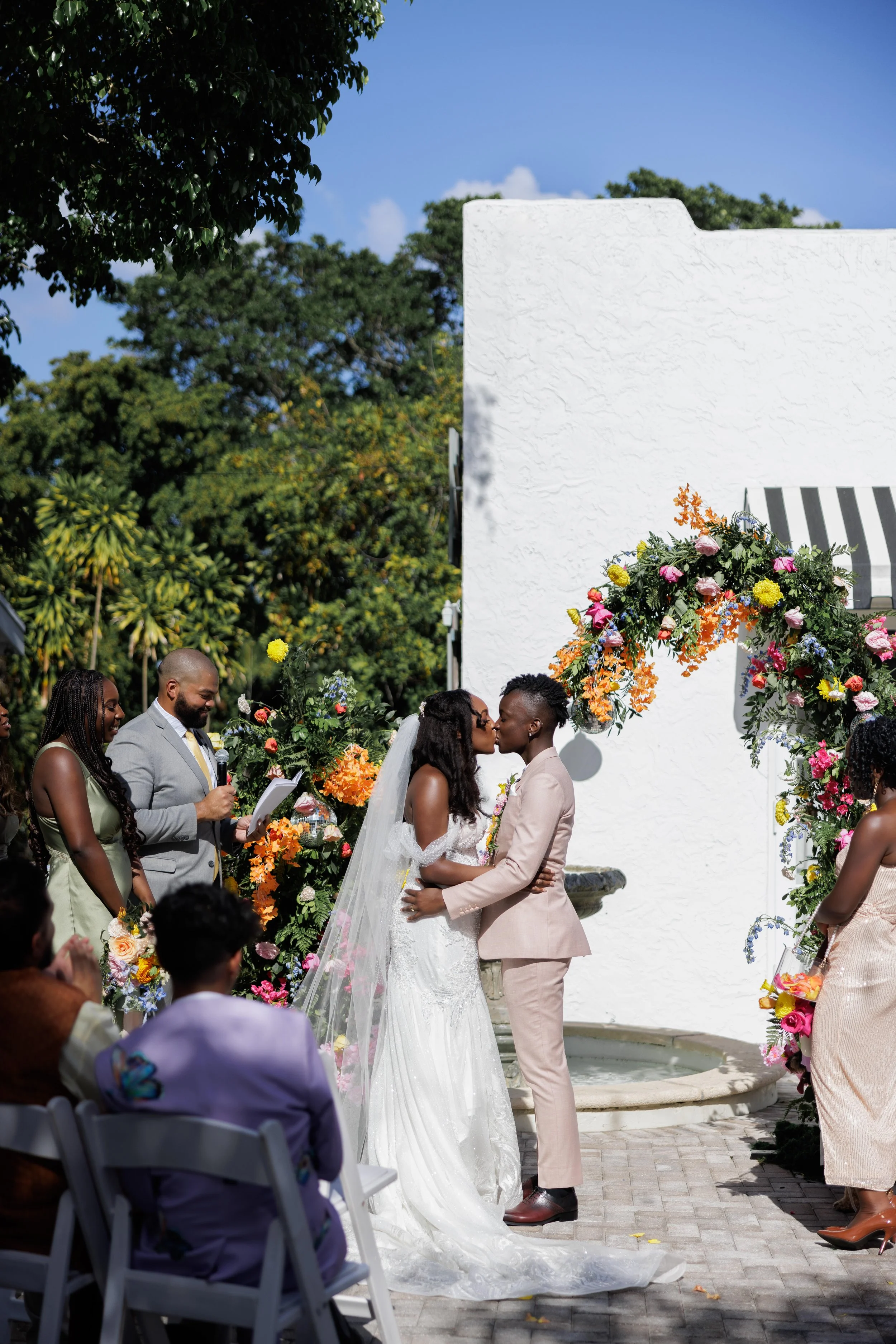 A couple sharing a kiss at their outdoor wedding ceremony, with guests and floral decorations surrounding them