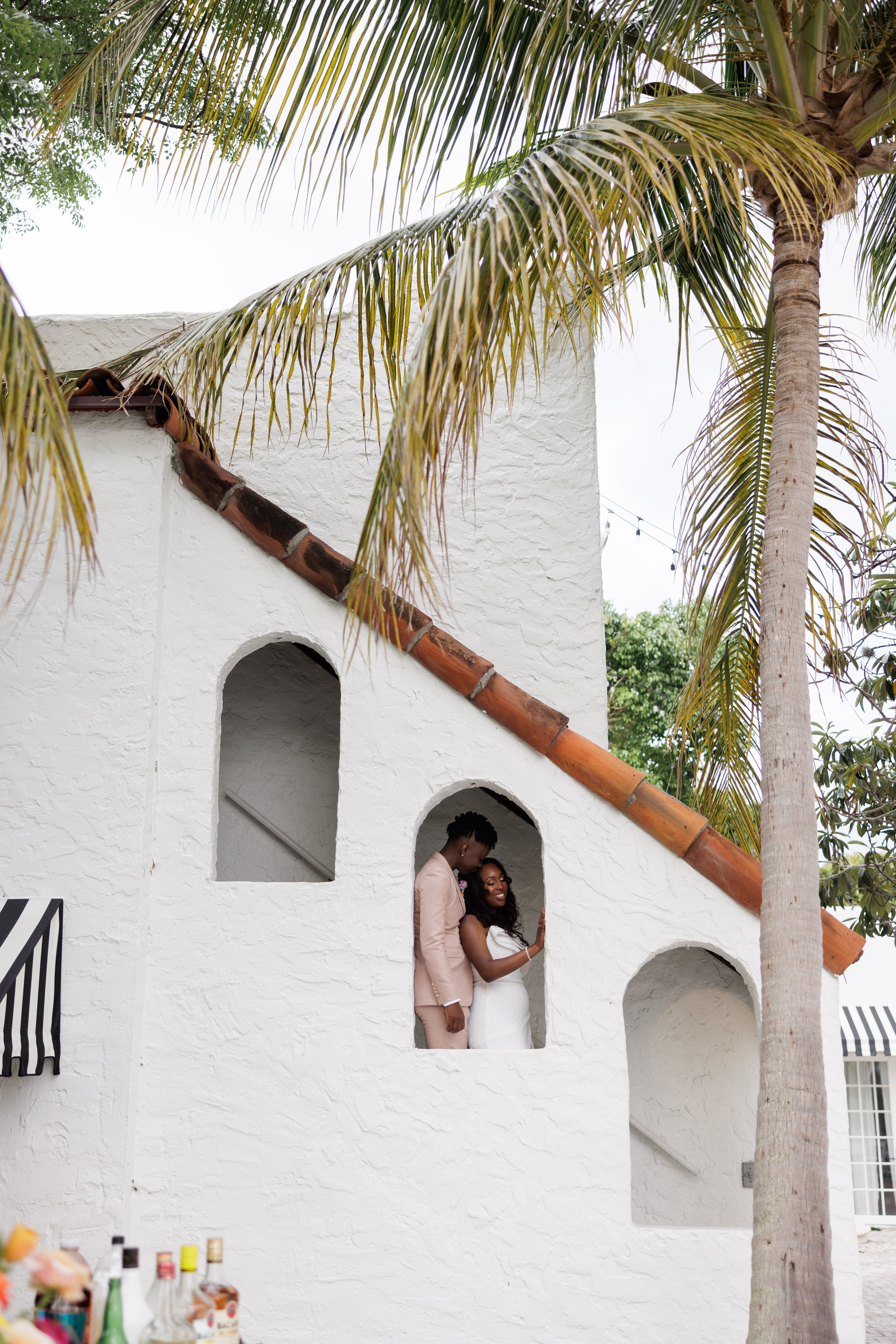 A couple dressed in formal attire standing on a white stucco staircase in a tropical setting with palm trees, smiling and sharing an intimate moment.
