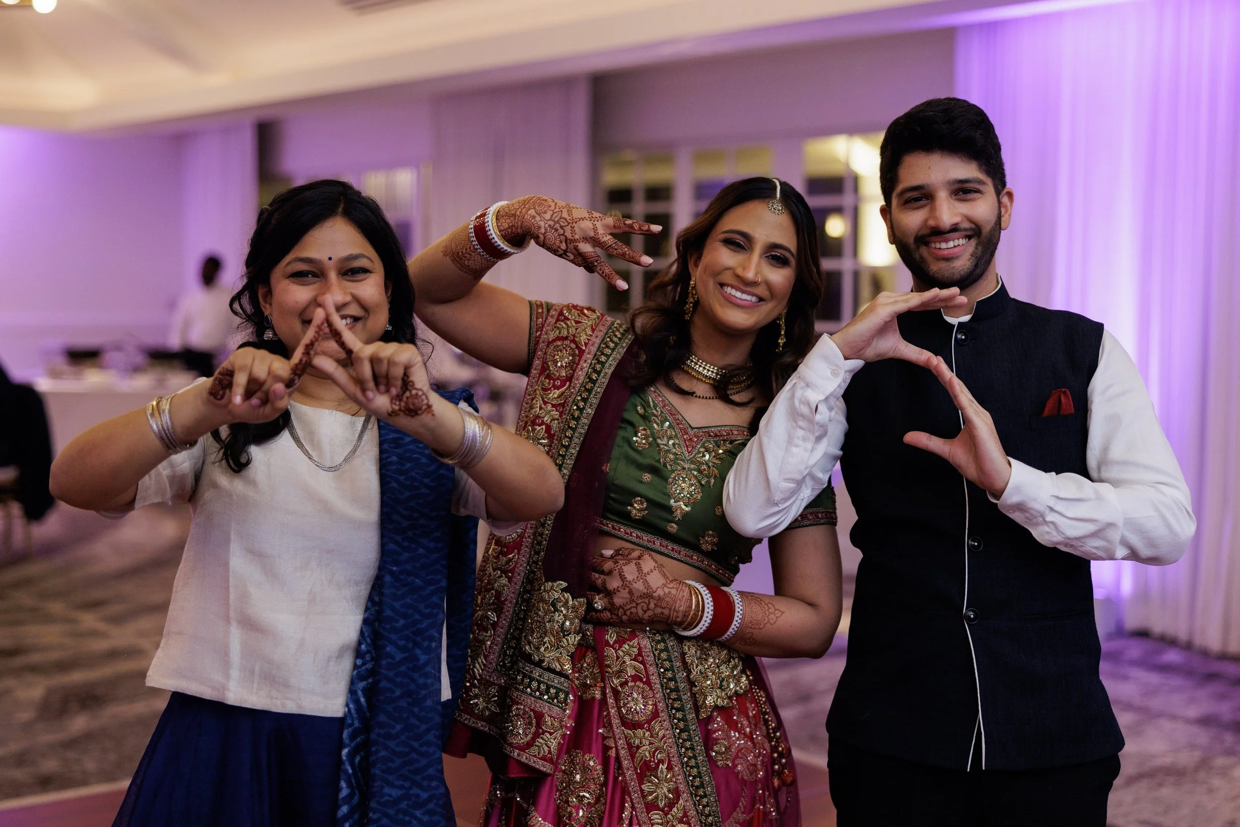 Three people at an Indian celebration, smiling and making heart shapes with their hands, indoors with purple lighting.
