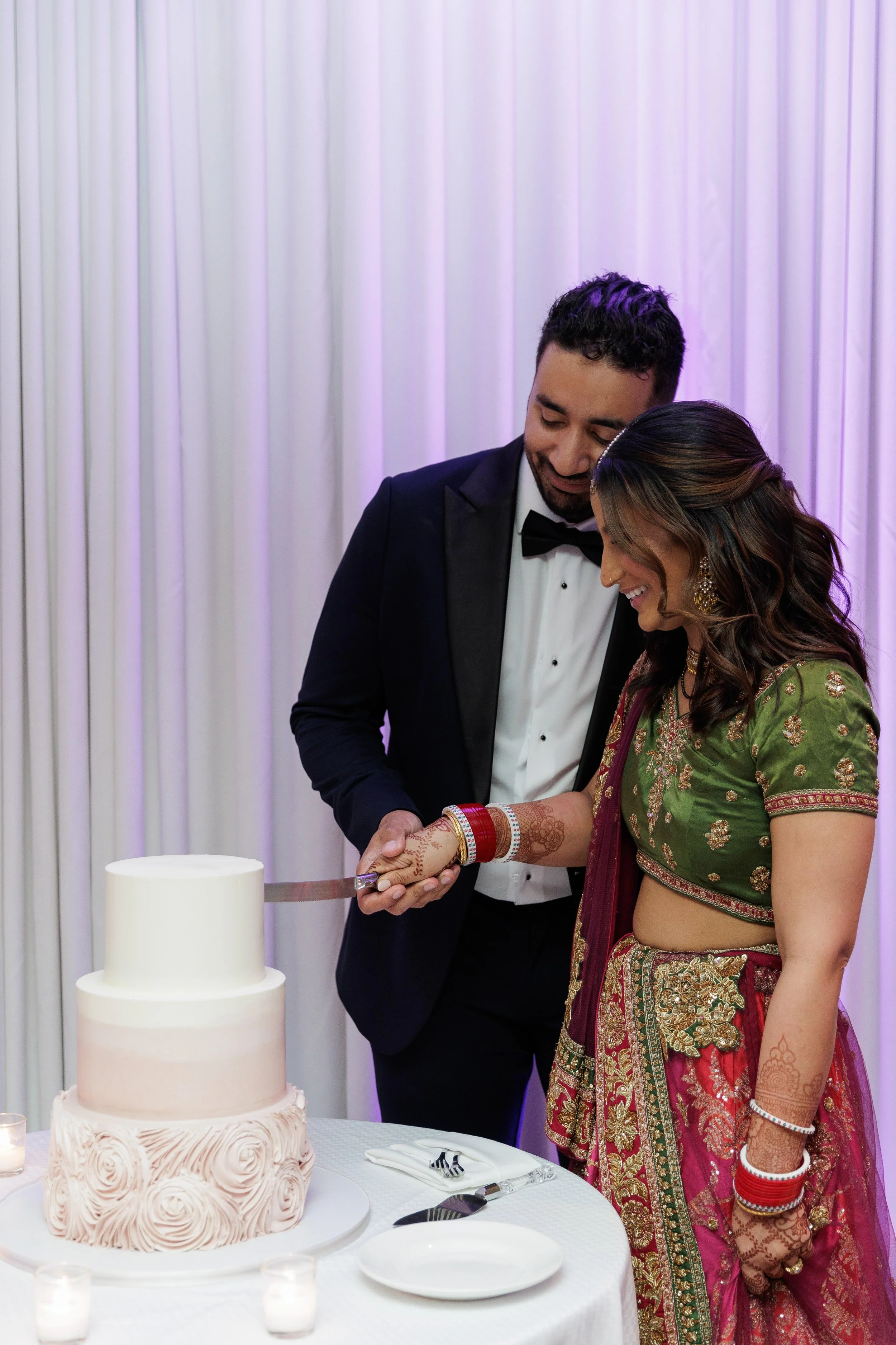 A couple cutting a wedding cake together, with the groom in a tuxedo and the bride in traditional Indian attire, smiling at each other. The cake is a three-tiered design on a white table with utensils and small candles around.