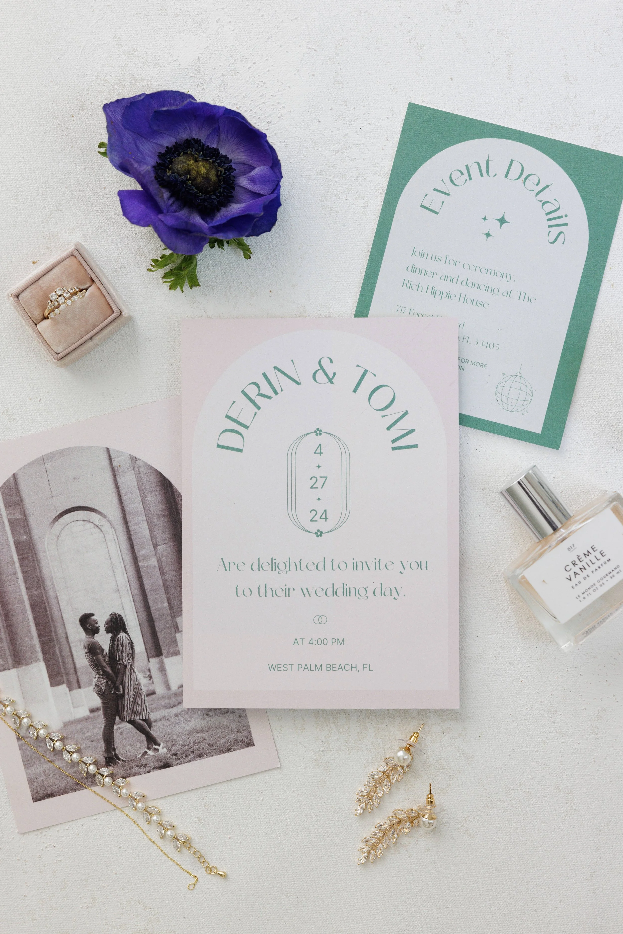Wedding invitation with a pink and mint green design, purple flower, pearl and gold jewelry, perfume bottle, and an engagement ring in a jewelry box on a white surface.