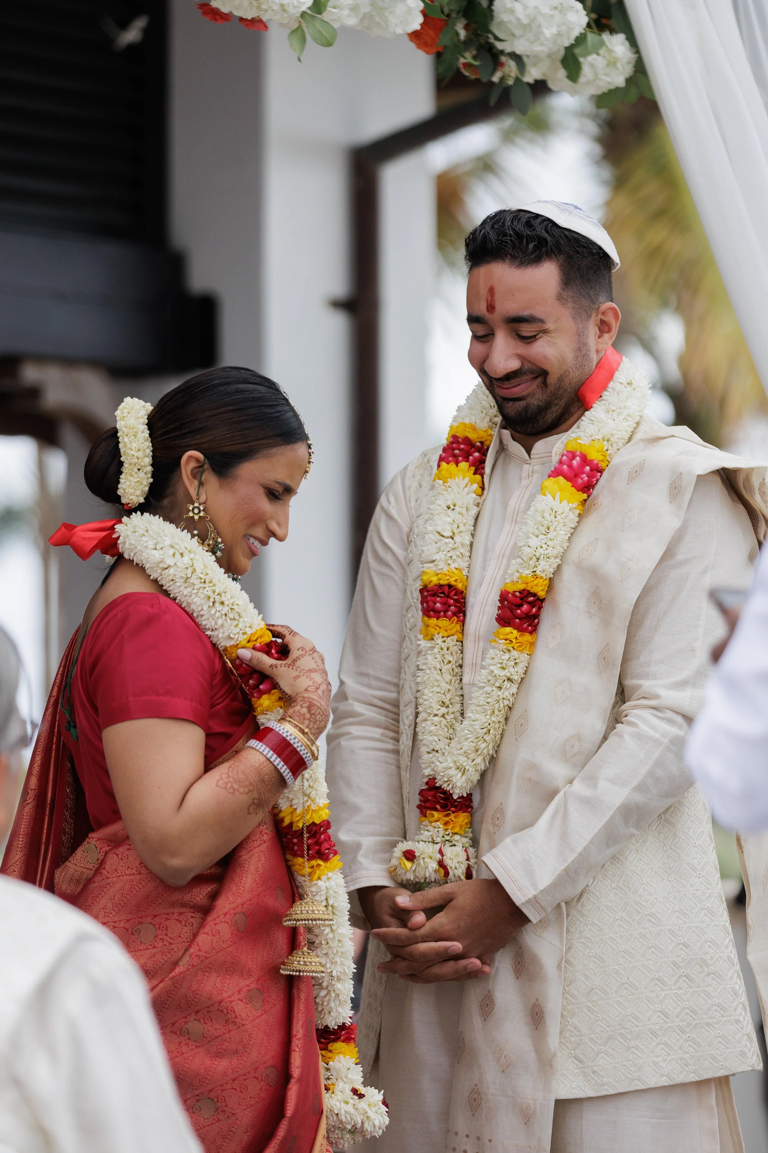 A couple participating in a traditional Indian wedding ceremony, wearing garlands of flowers, standing close together, smiling with their eyes closed.
