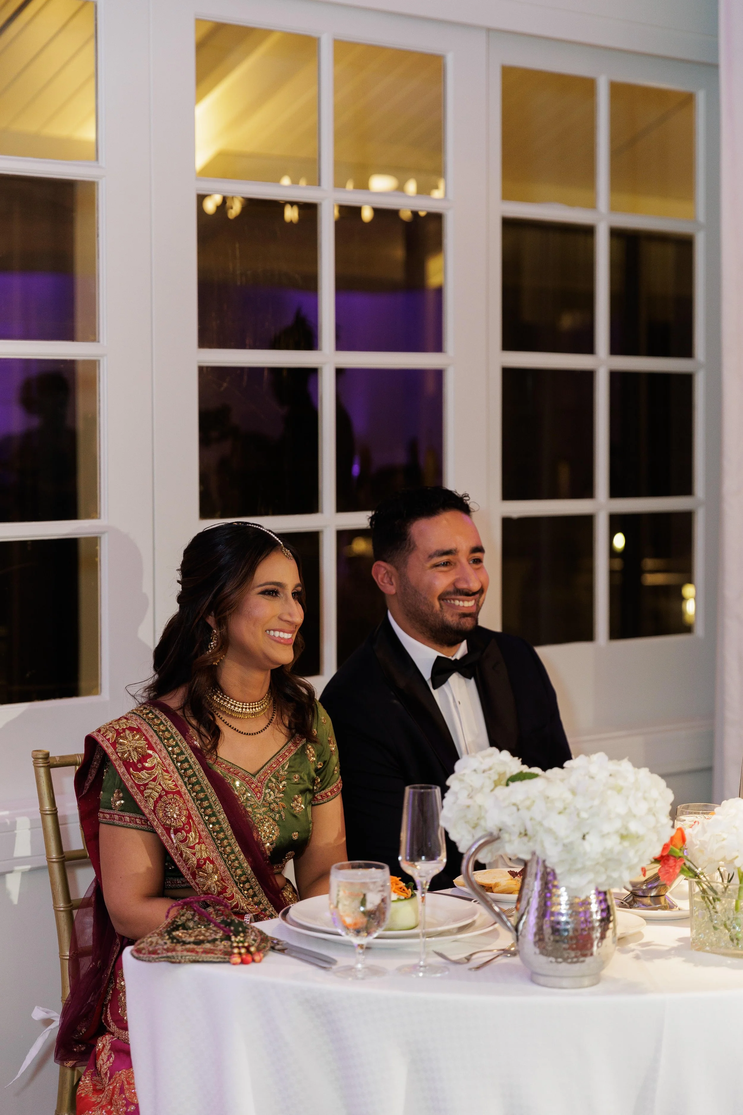 Two people, a woman in a traditional South Asian outfit and a man in a tuxedo, sitting at a decorated wedding reception table with white flowers and elegant tableware, smiling.