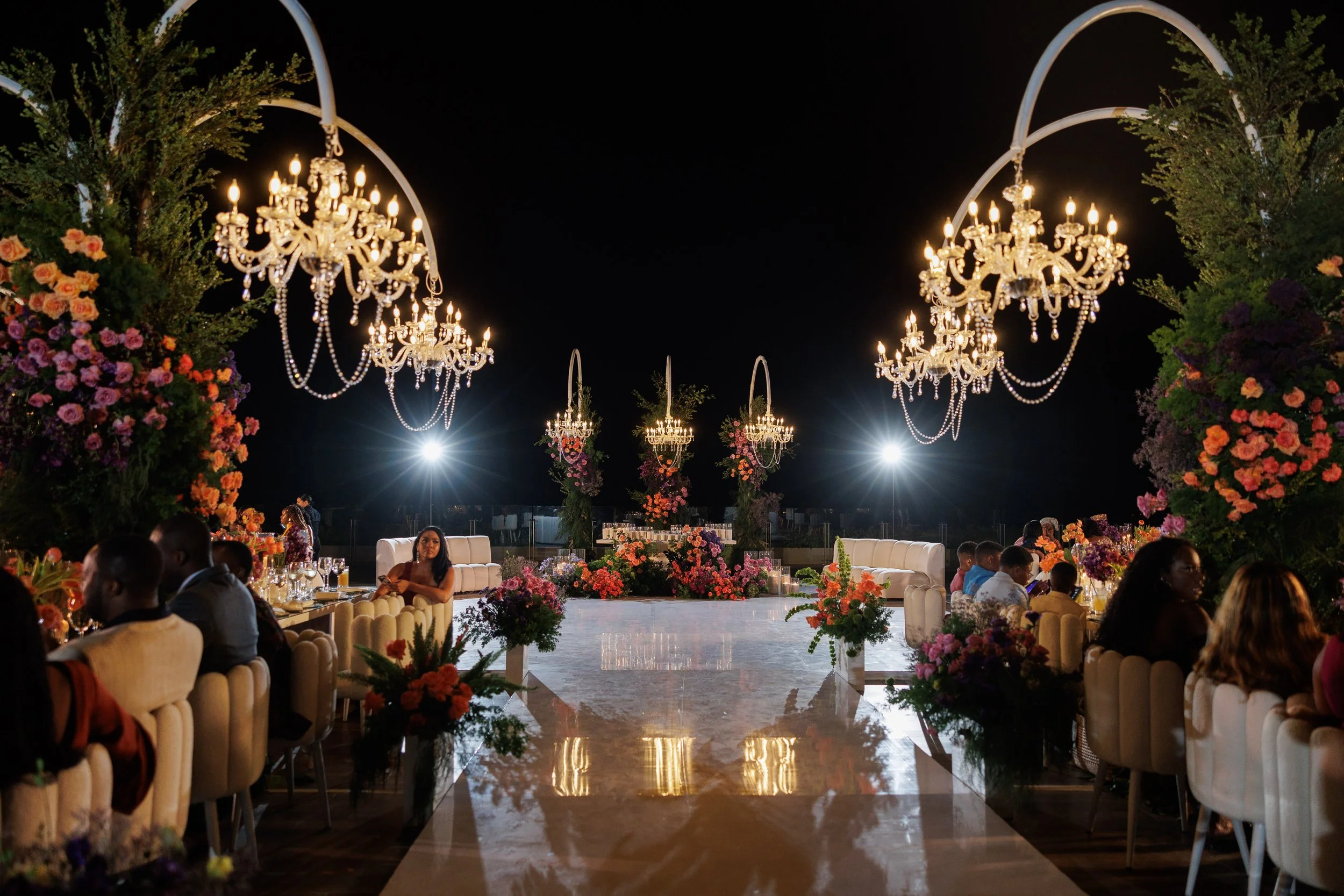 Elegant outdoor event at night with chandeliers, floral arrangements, and seated guests along a decorated aisle.