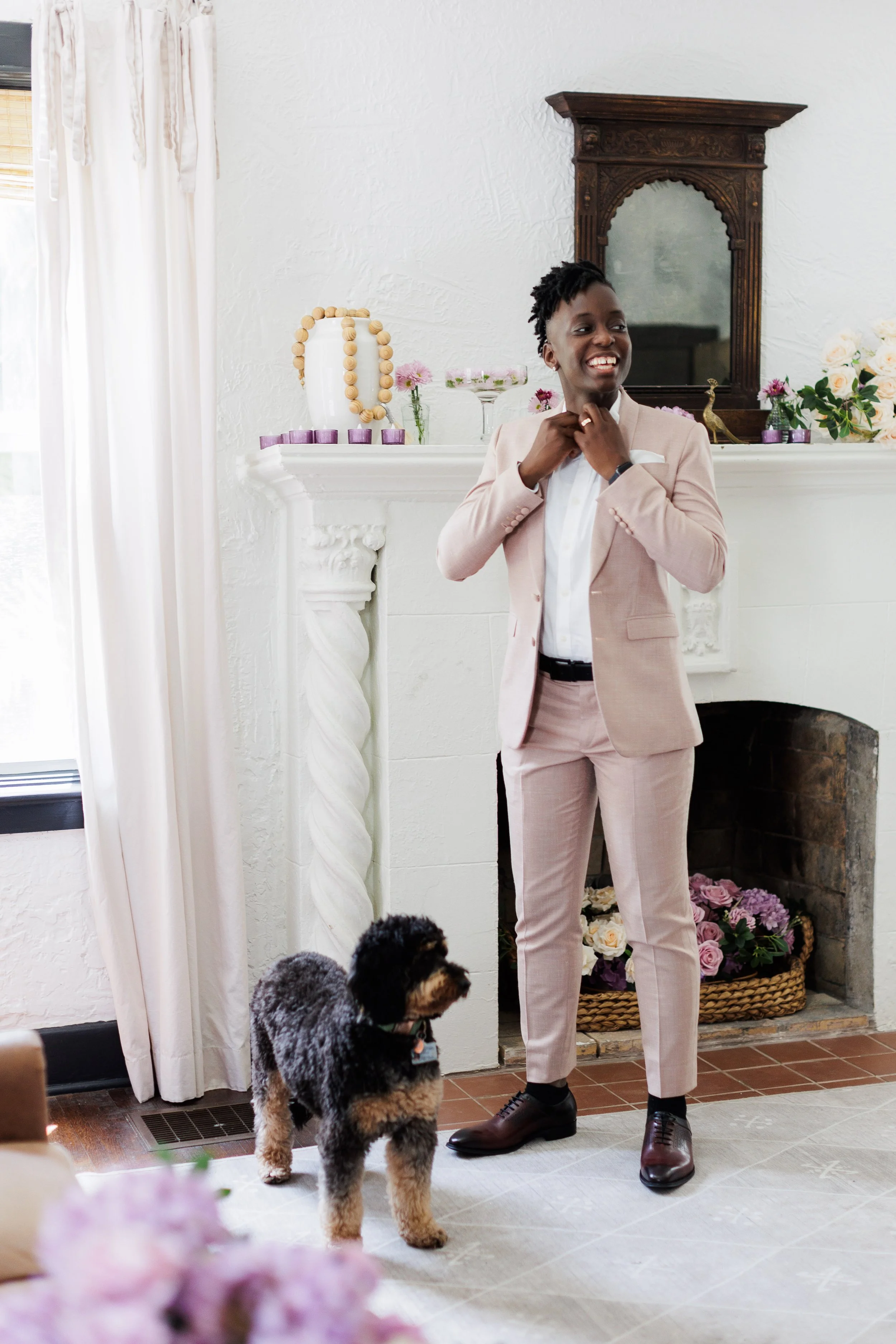A woman in a pink suit smiling and adjusting her collar while standing next to a dog inside a living room with a fireplace and decorative mantle.