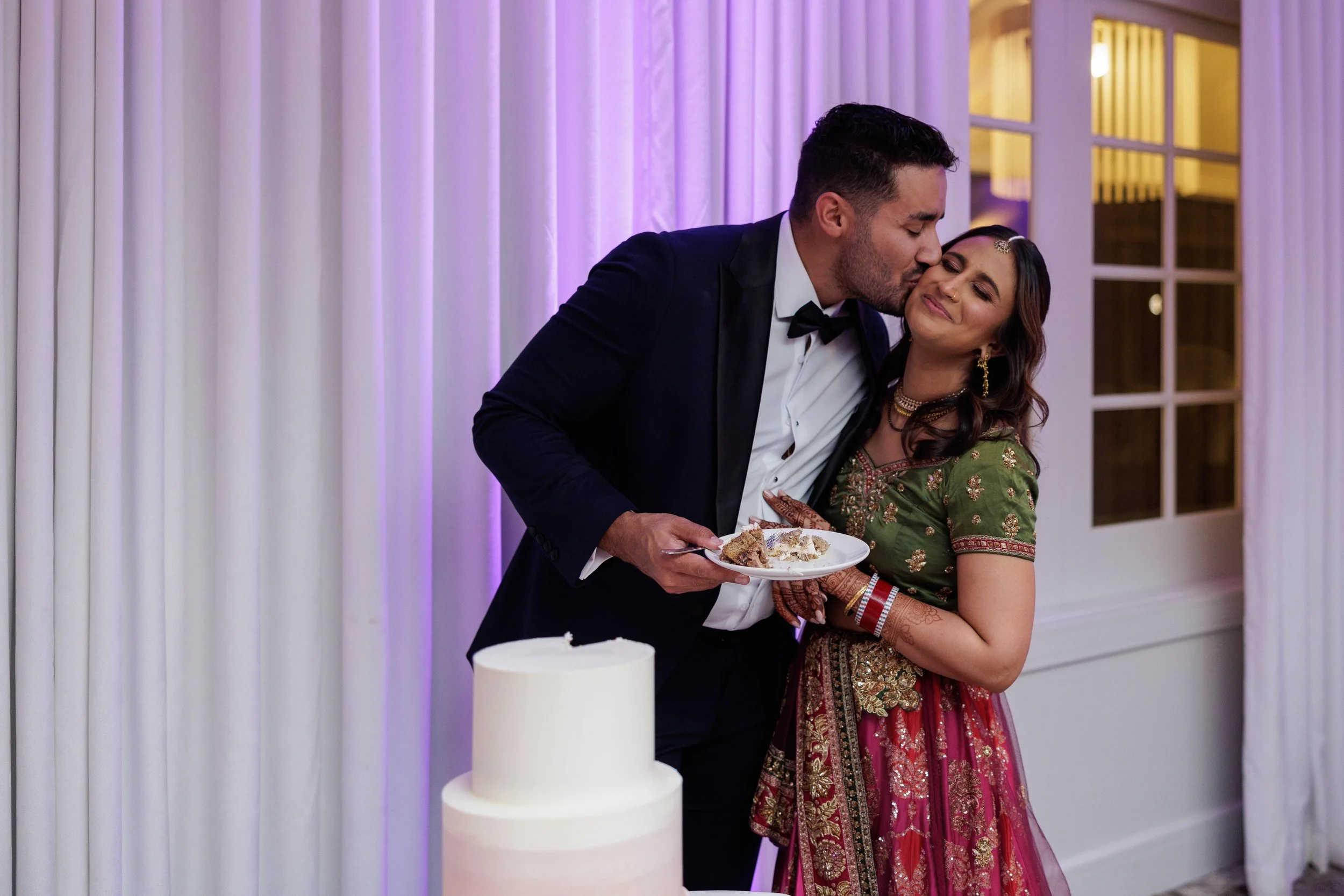 A man in a tuxedo is kissing a woman in a traditional Indian dress on the cheek at a celebration. They are standing next to a white cake, and the woman is smiling with her eyes closed. The background has purple lights and white curtains.
