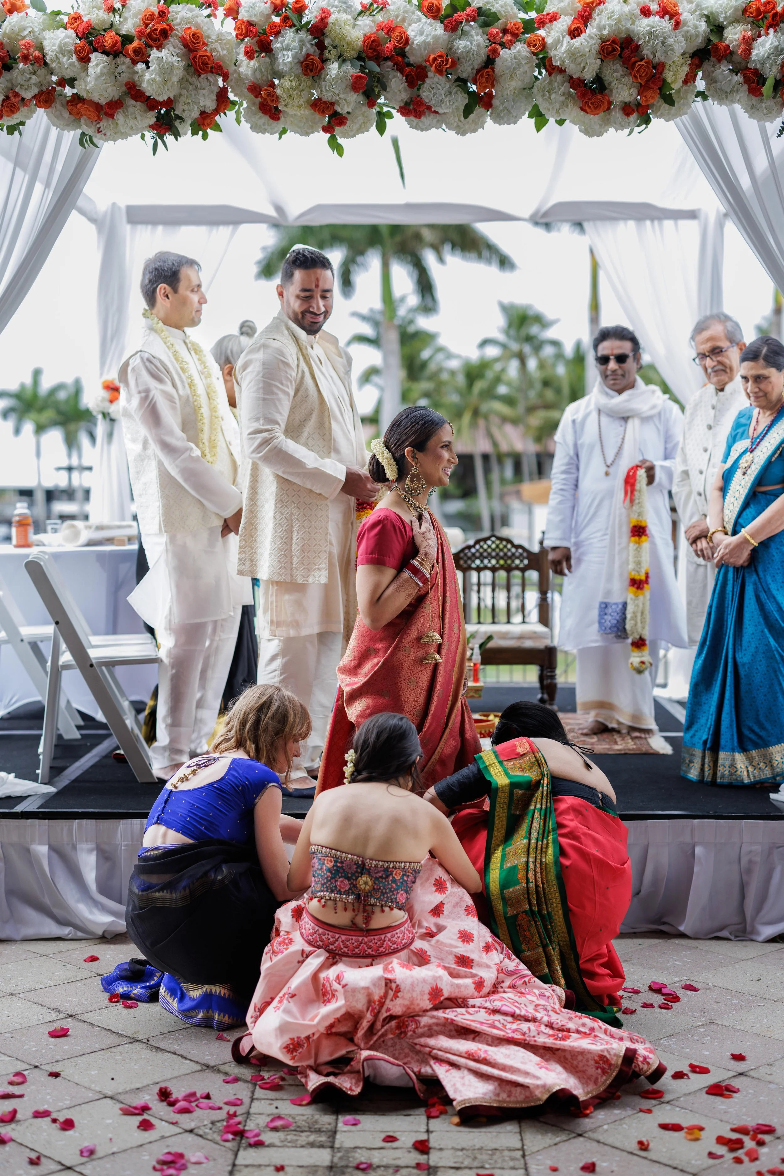 A traditional Indian wedding ceremony with a bride in a red sari, seated on the ground, while women in colorful saris perform rituals. The groom and other men in traditional white attire with flower garlands observe, under a decorated canopy with whi