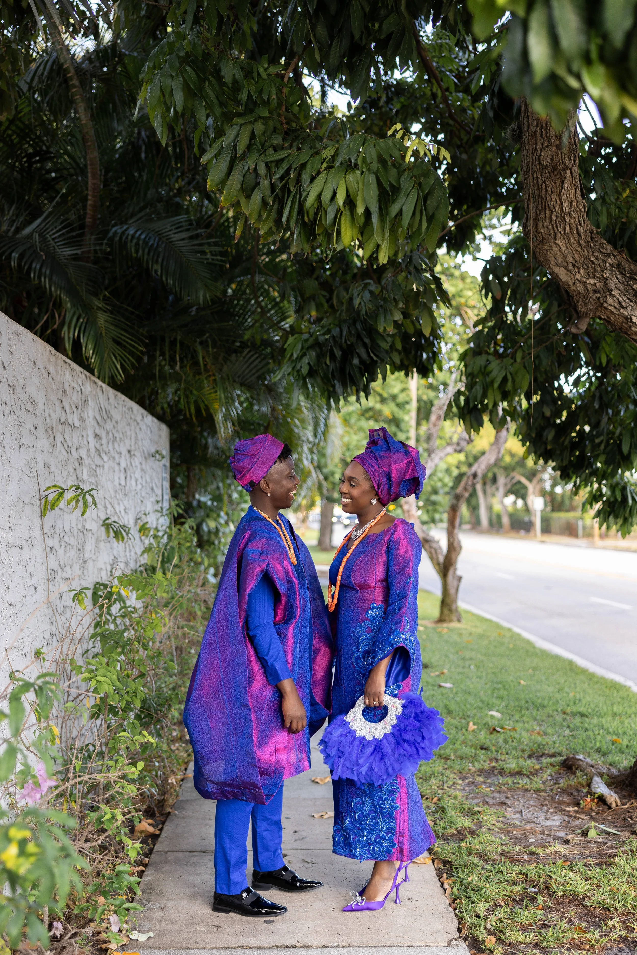 Two women standing on a sidewalk, wearing traditional Nigerian attire in purple and blue, smiling at each other under a leafy tree.