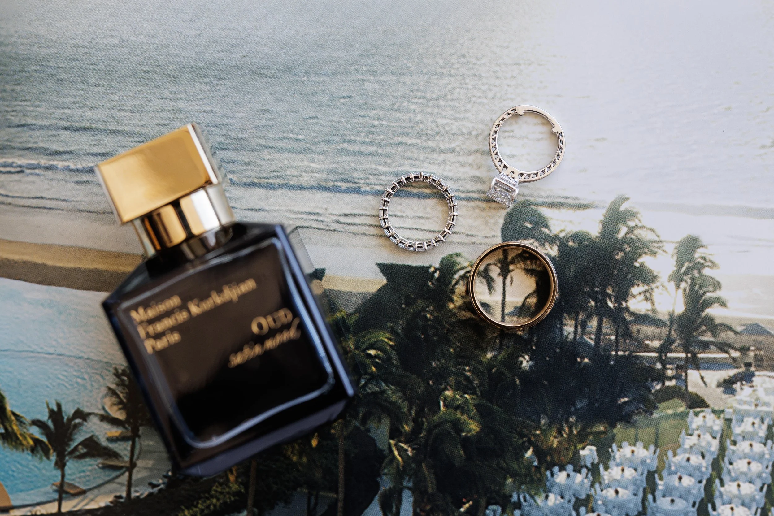 A bottle of Maison Francis Kurkdjian Paris Oud perfume, two silver rings, and a gold ring with a reflection of palm trees, all placed on a beach scene background.