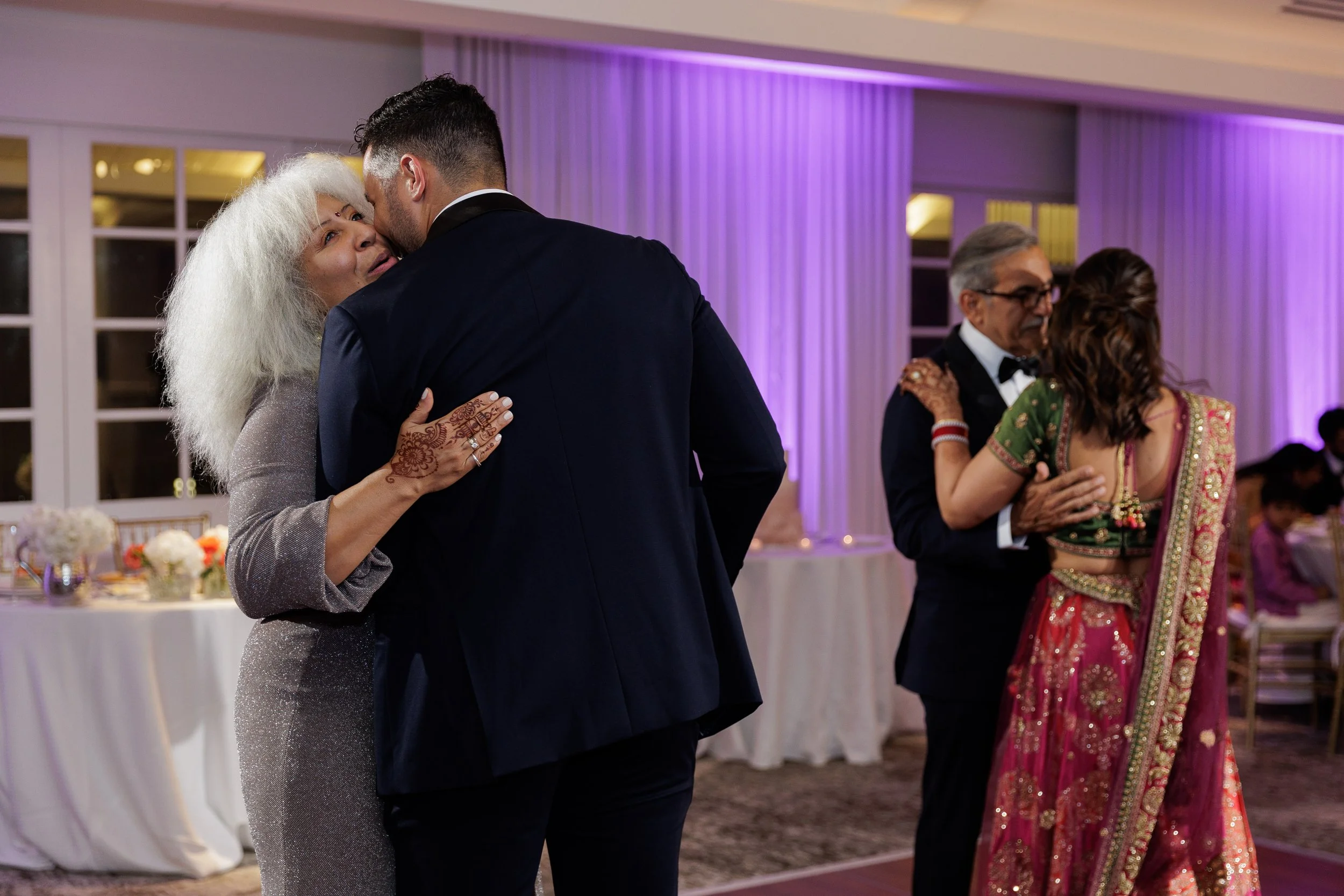 Older woman with curly white hair and henna on her hand dancing closely with young man in a tuxedo, in a decorated event hall with purple lighting.