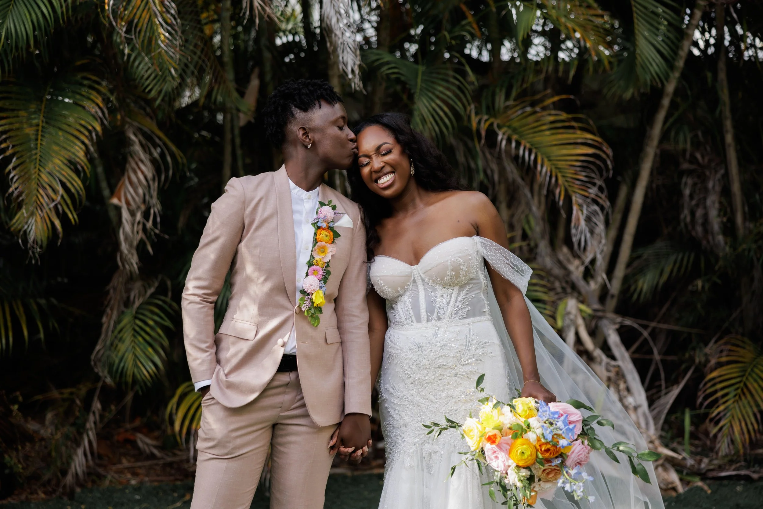 A bride and groom holding hands during their wedding, with the groom kissing the bride's forehead and the bride smiling, surrounded by tropical plants.