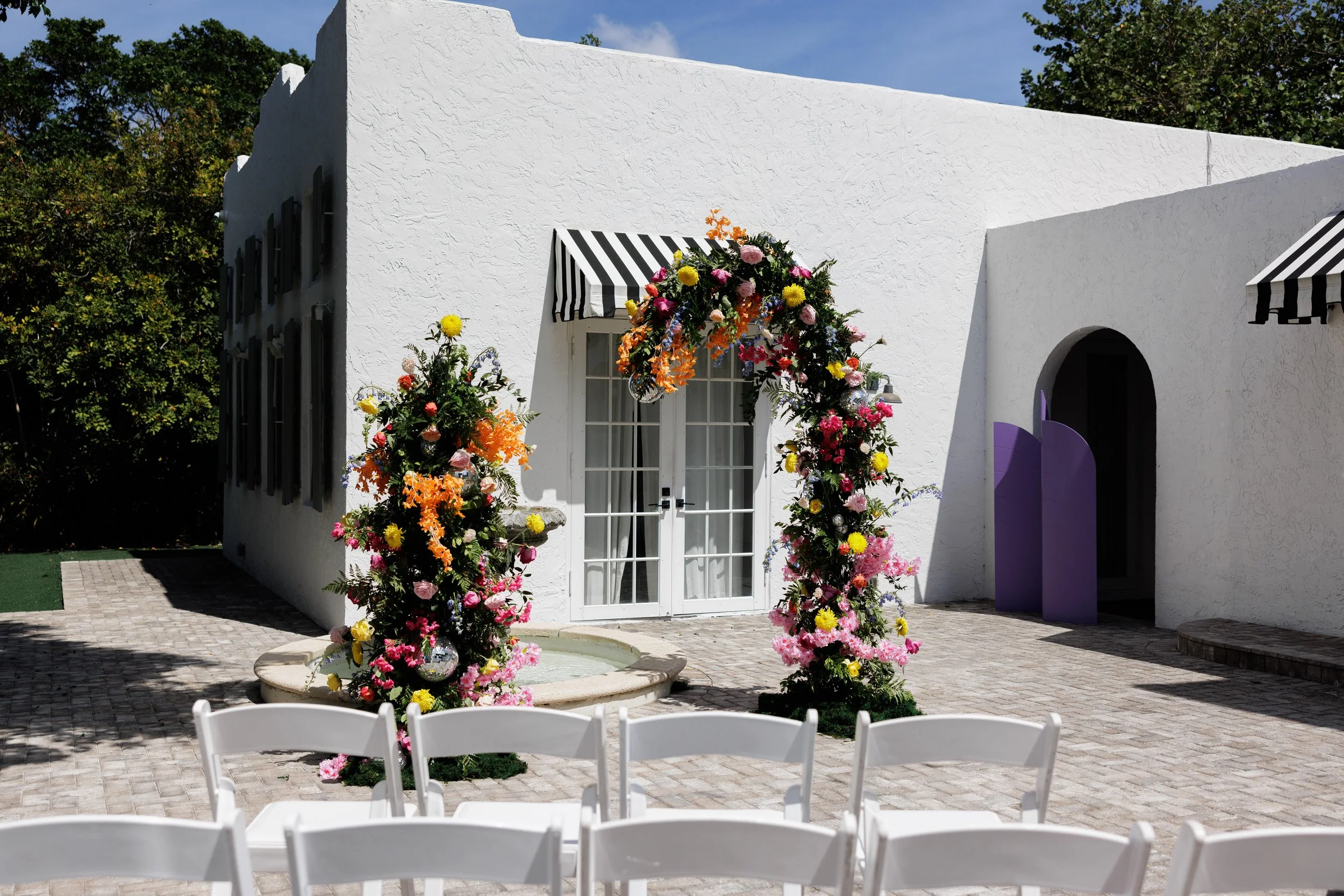 Decorated white building exterior with floral arch and chairs, indicating a wedding setup.