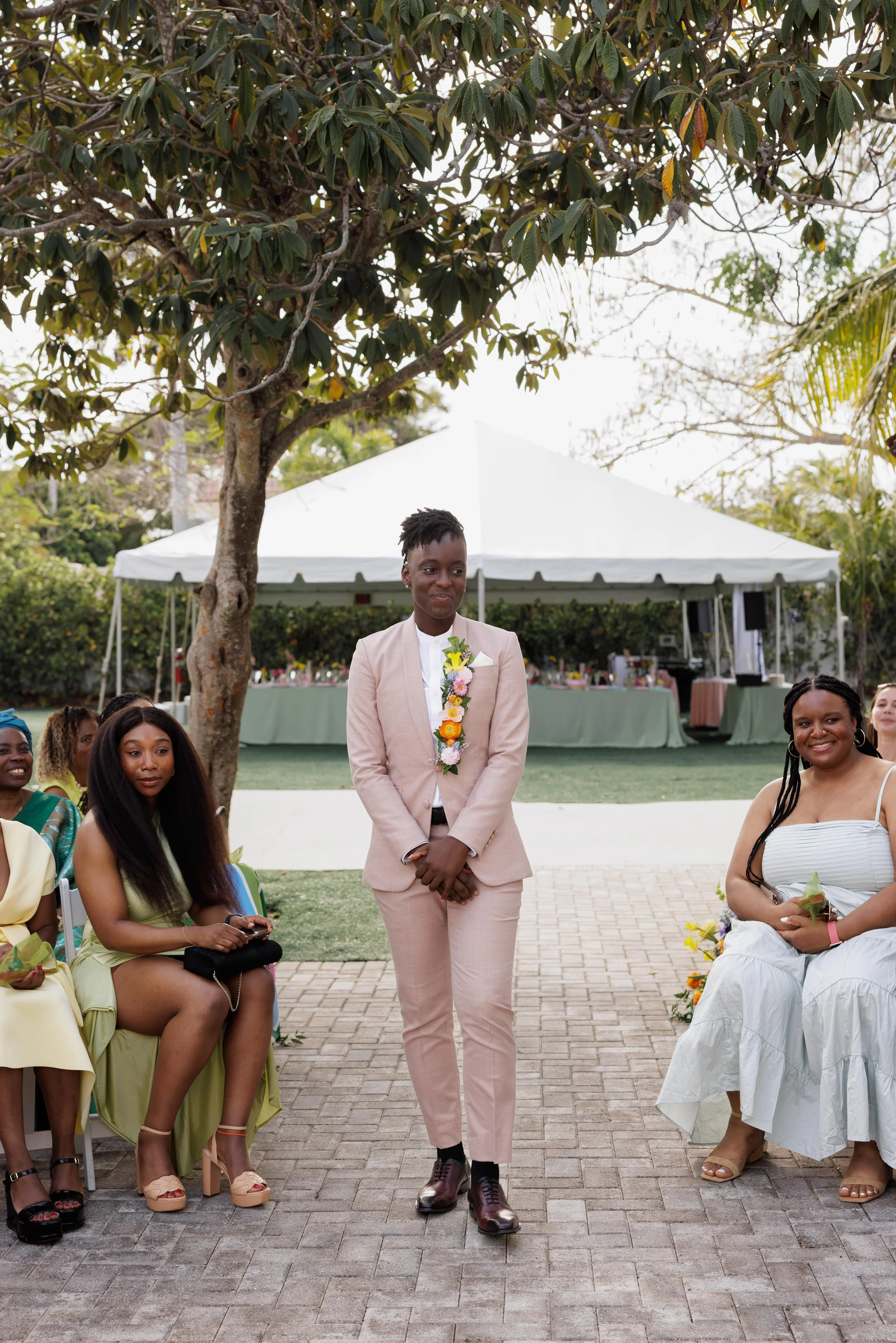 A woman in a pink suit and floral garland standing outdoors during a celebration, with other women seated nearby and a large white tent in the background.
