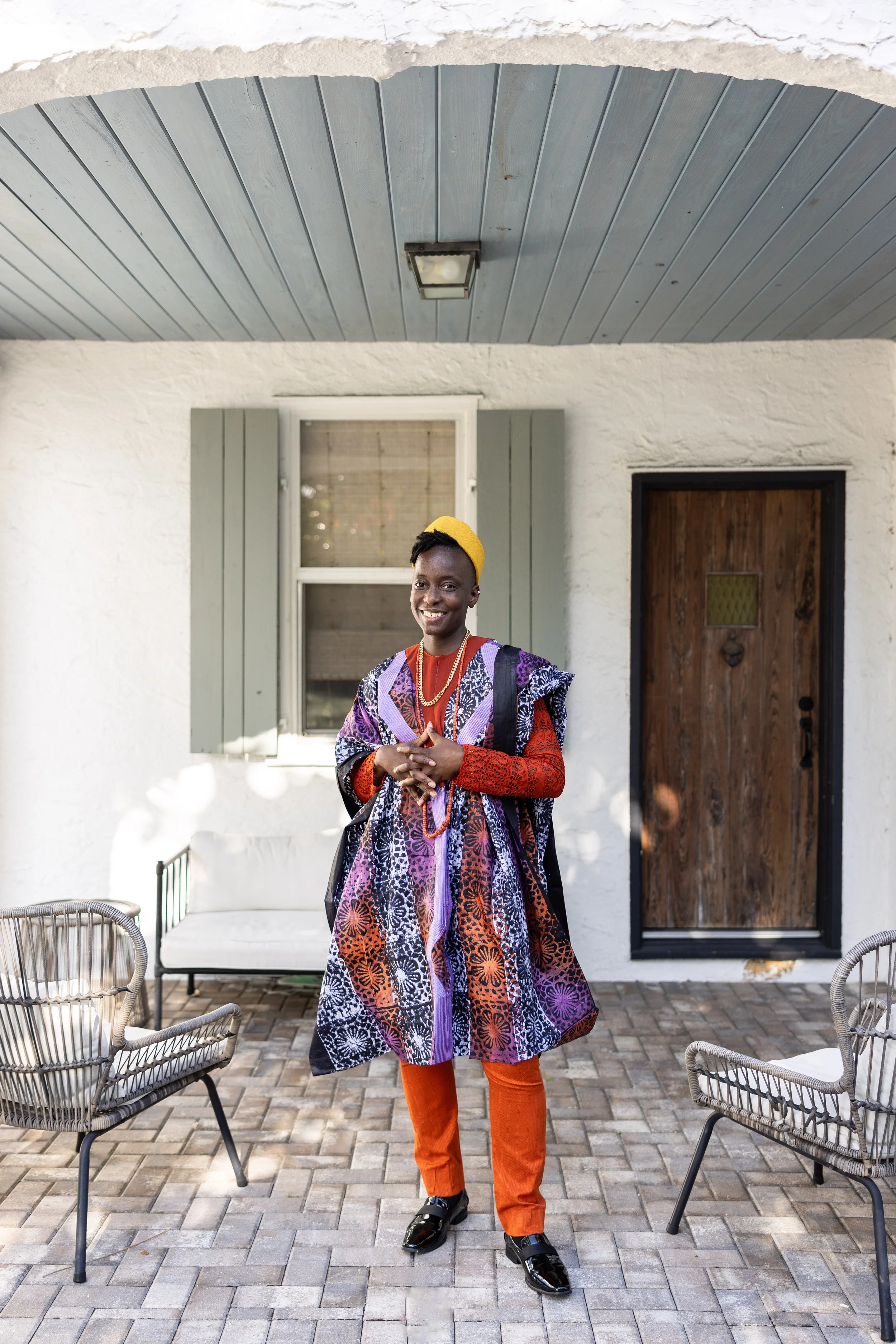 A smiling woman standing outdoors on a patio in front of a white stucco house with gray window shutters and a wooden door. She is wearing a colorful, patterned dress, orange pants, black shoes, and a yellow headband, with a black backpack on her shou